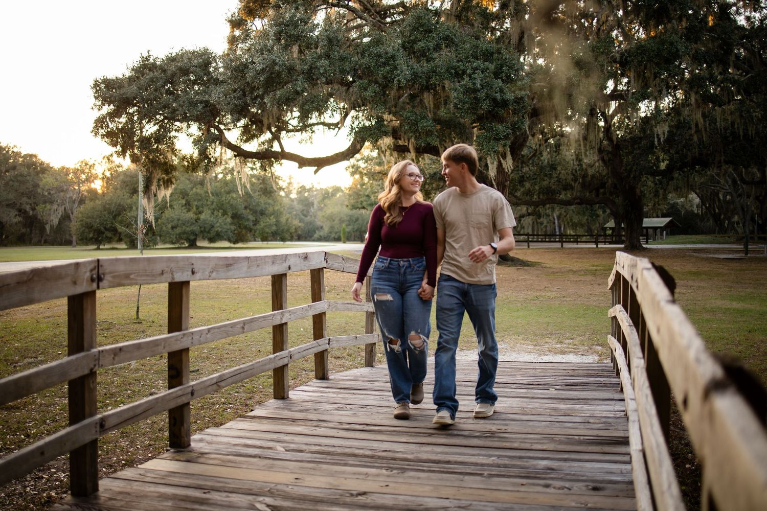 A couple walks hand in hand across a wooden bridge in a park at sunset.