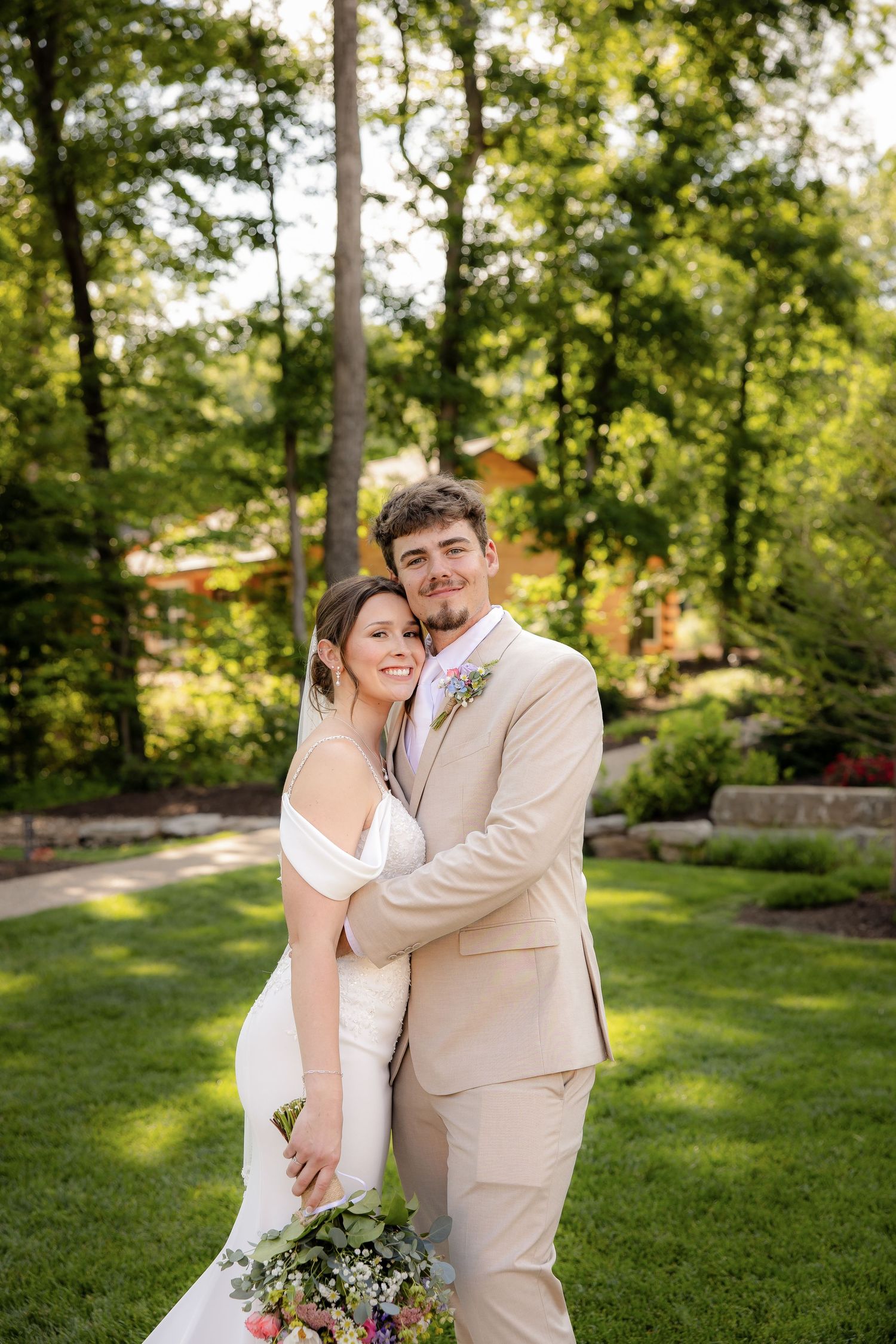 A bride and groom embrace outdoors on green grass, she holds a floral bouquet, he wears a tan suit with boutonniere.