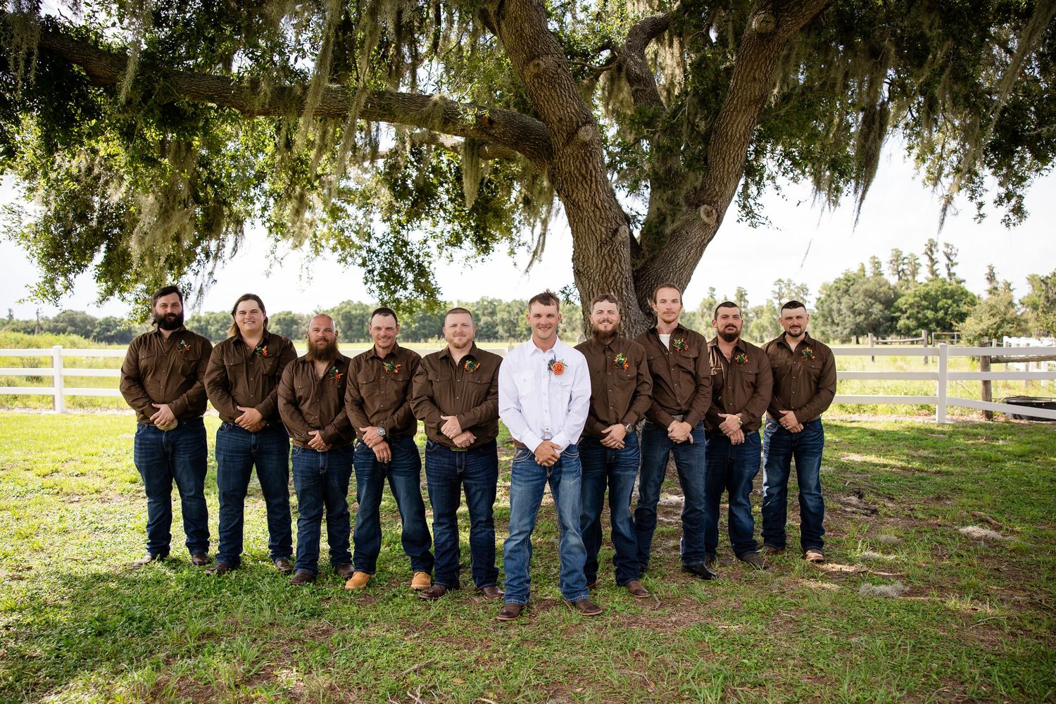 Groom in white shirt with groomsmen in brown shirts and jeans posing under a large oak tree on a farm.