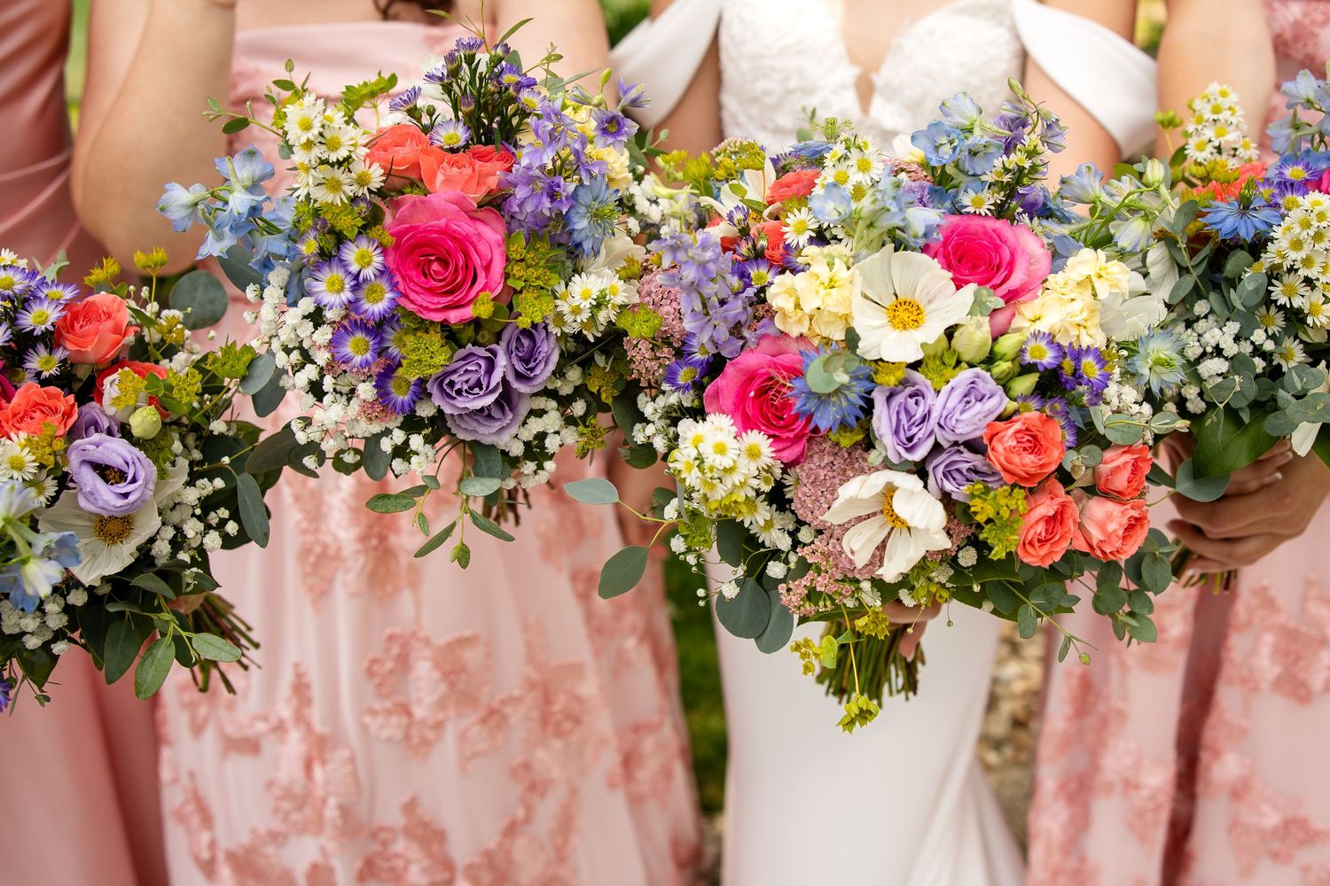 Bridesmaids in pink dresses holding colorful wedding bouquets with roses, wildflowers, and greenery alongside the bride.