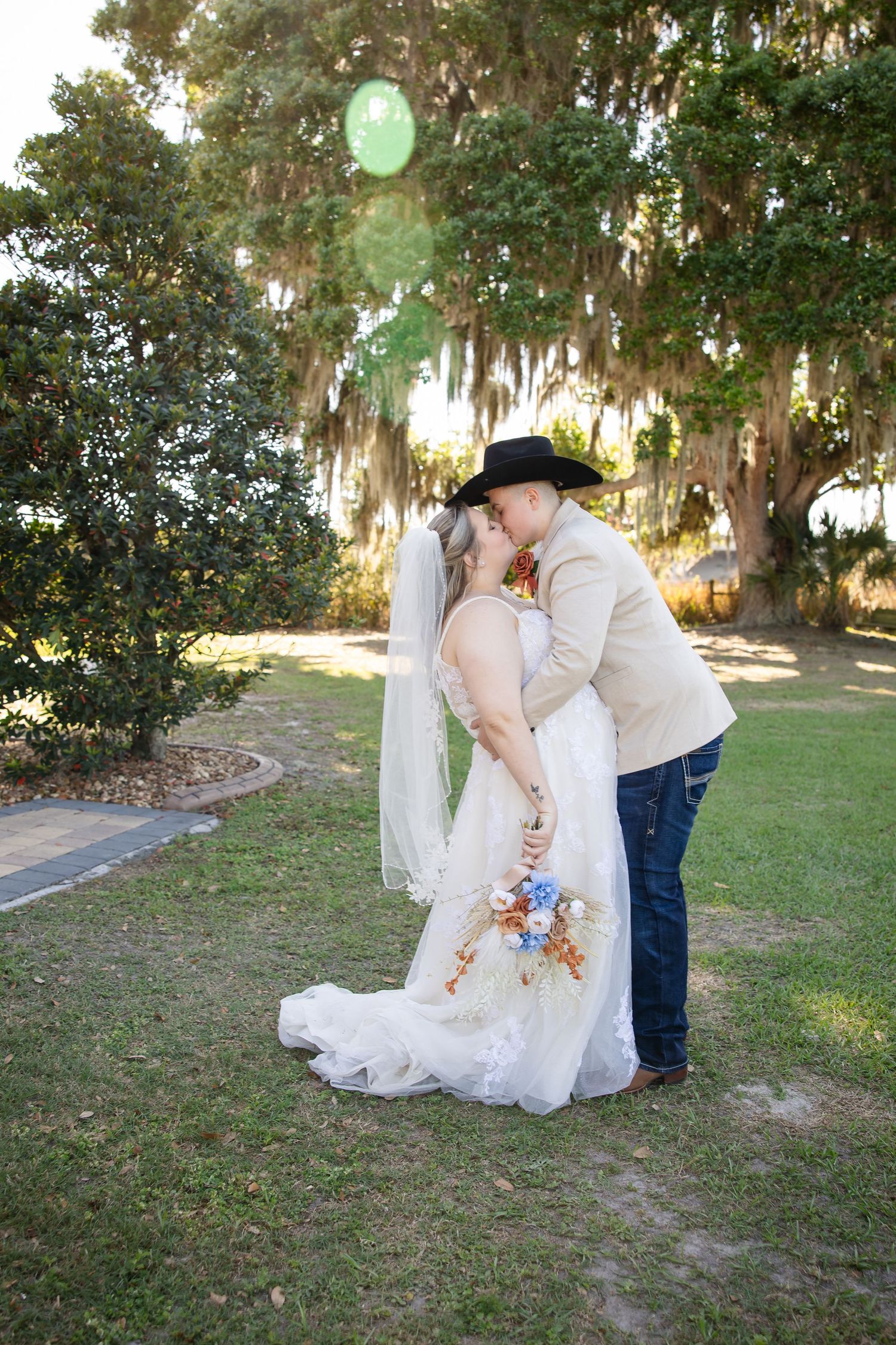 Couple sharing a kiss outdoors, bride in white gown with veil, partner in cowboy hat and jeans, surrounded by moss-draped trees.