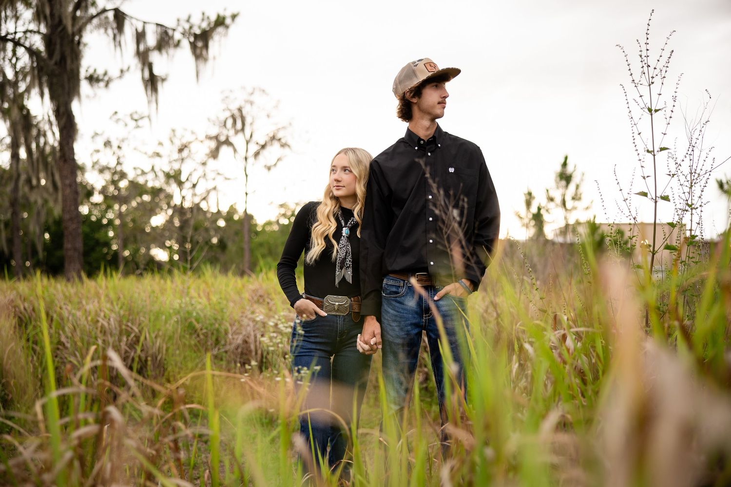 Couple holding hands in tall grass field with Spanish moss trees, wearing black shirts and jeans in outdoor engagement photo.