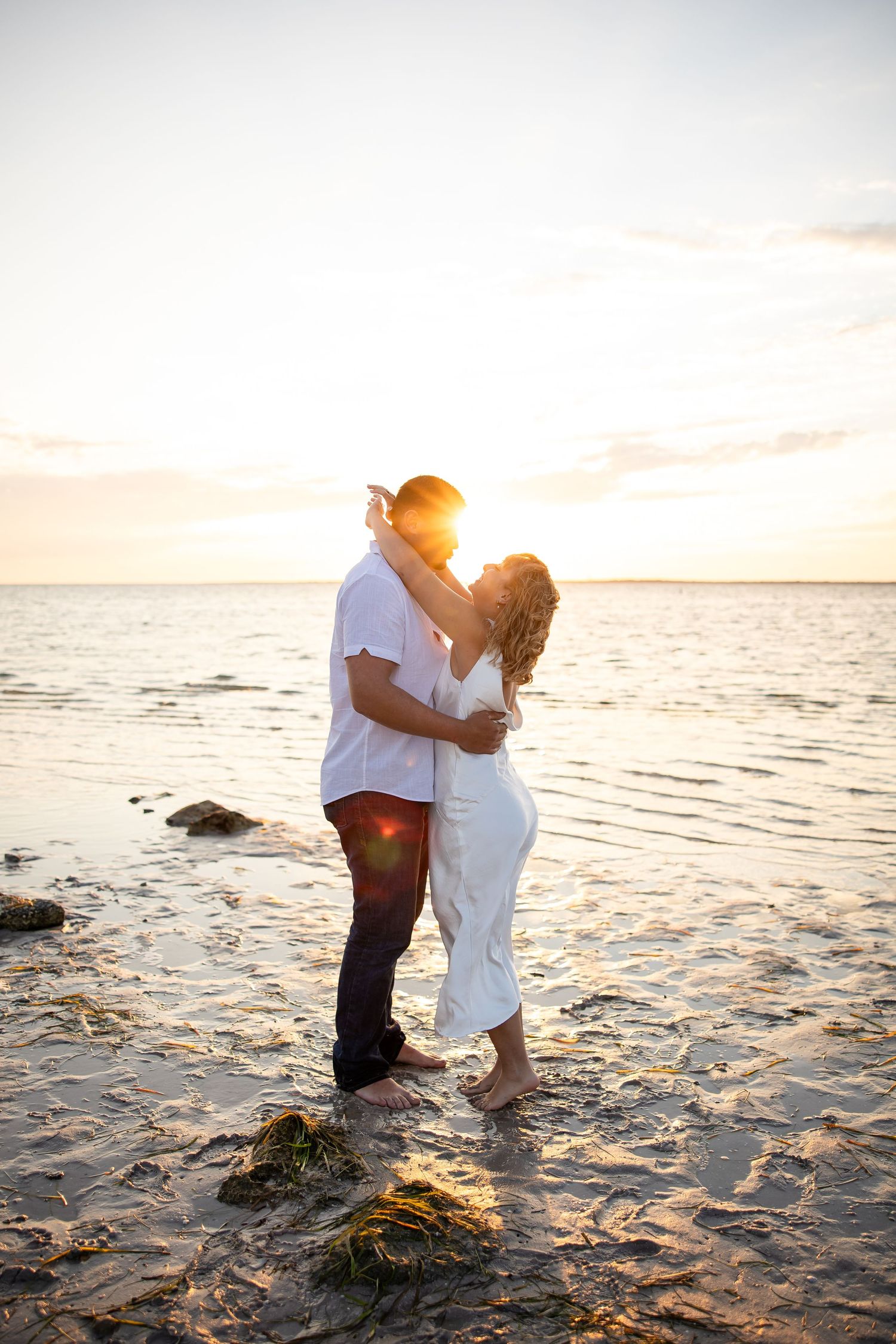 Couple embracing and kissing on a sandy beach at golden sunset with ocean water reflecting warm light.