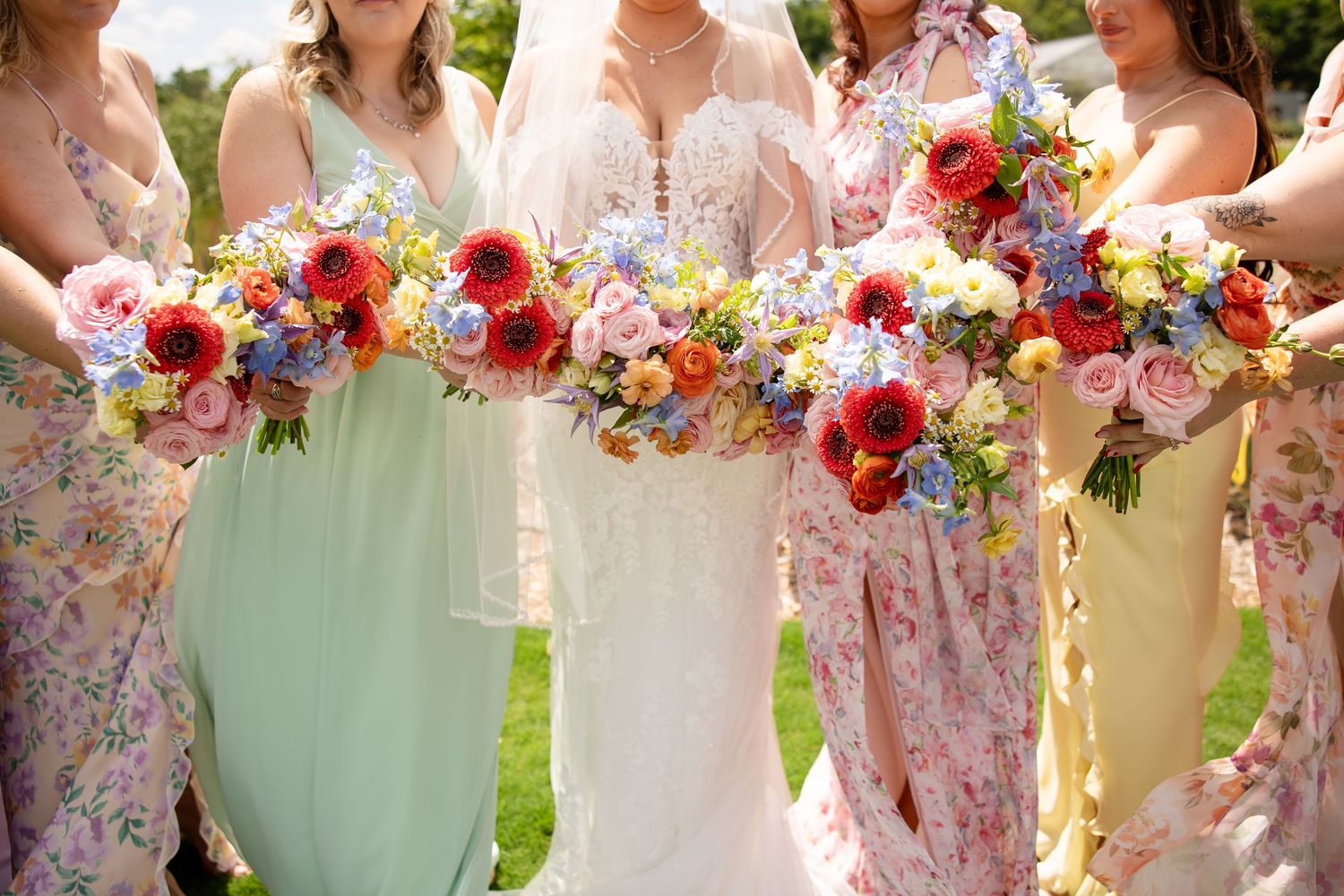 Bride in white lace gown with bridesmaids in floral and pastel dresses holding vibrant colorful wedding bouquets outdoors.