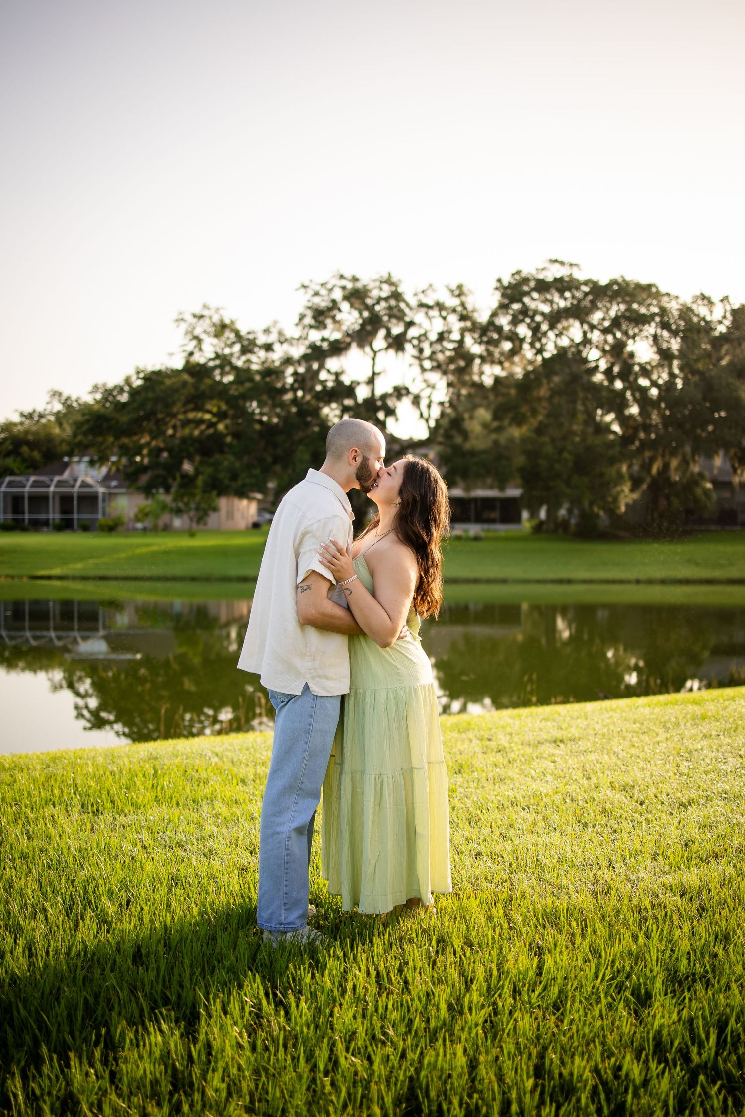 A romantic couple shares a tender moment at sunset by a tranquil pond on a grassy lawn.