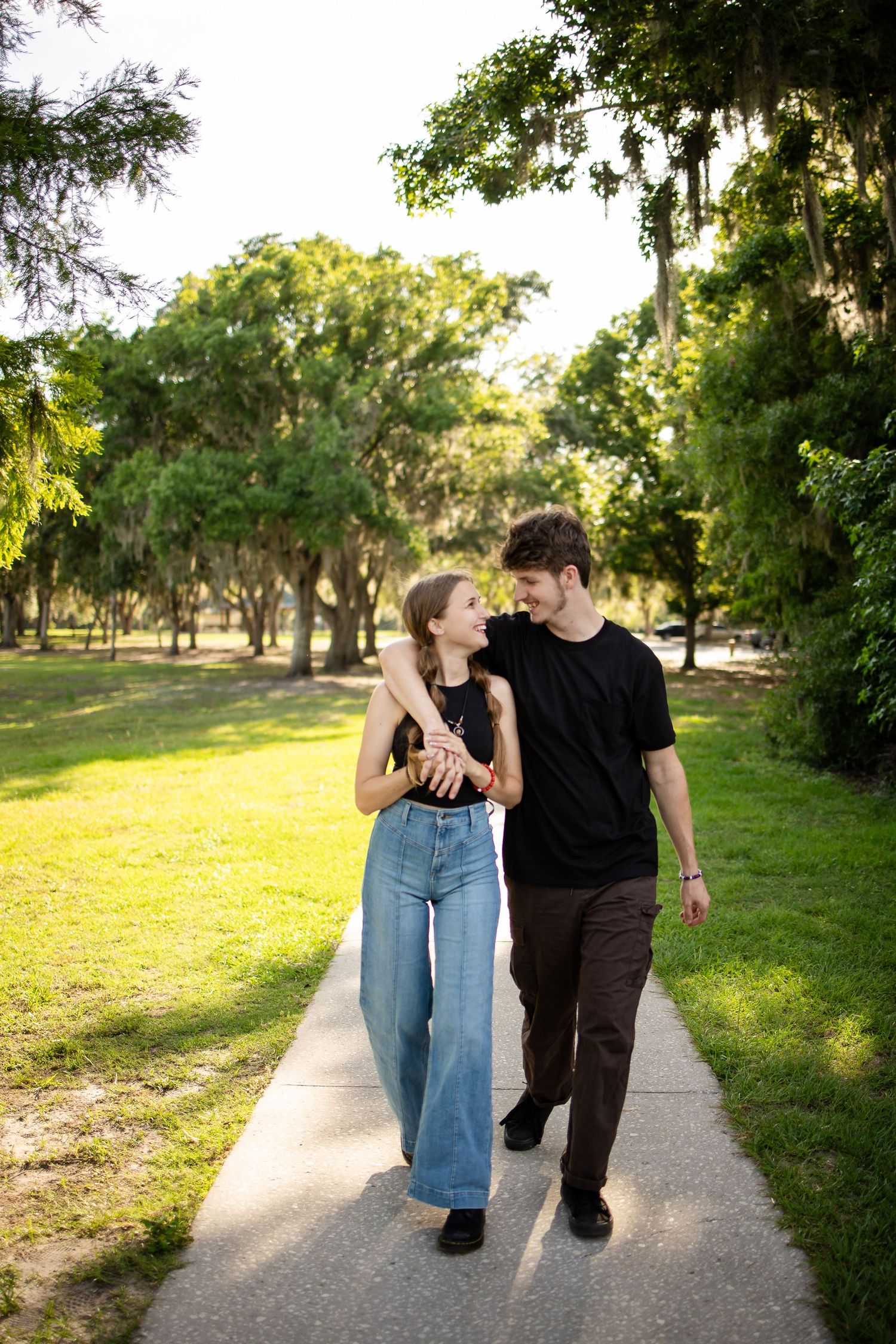 A couple walks together on a sunlit path through a lush park with Spanish moss hanging from trees above them.