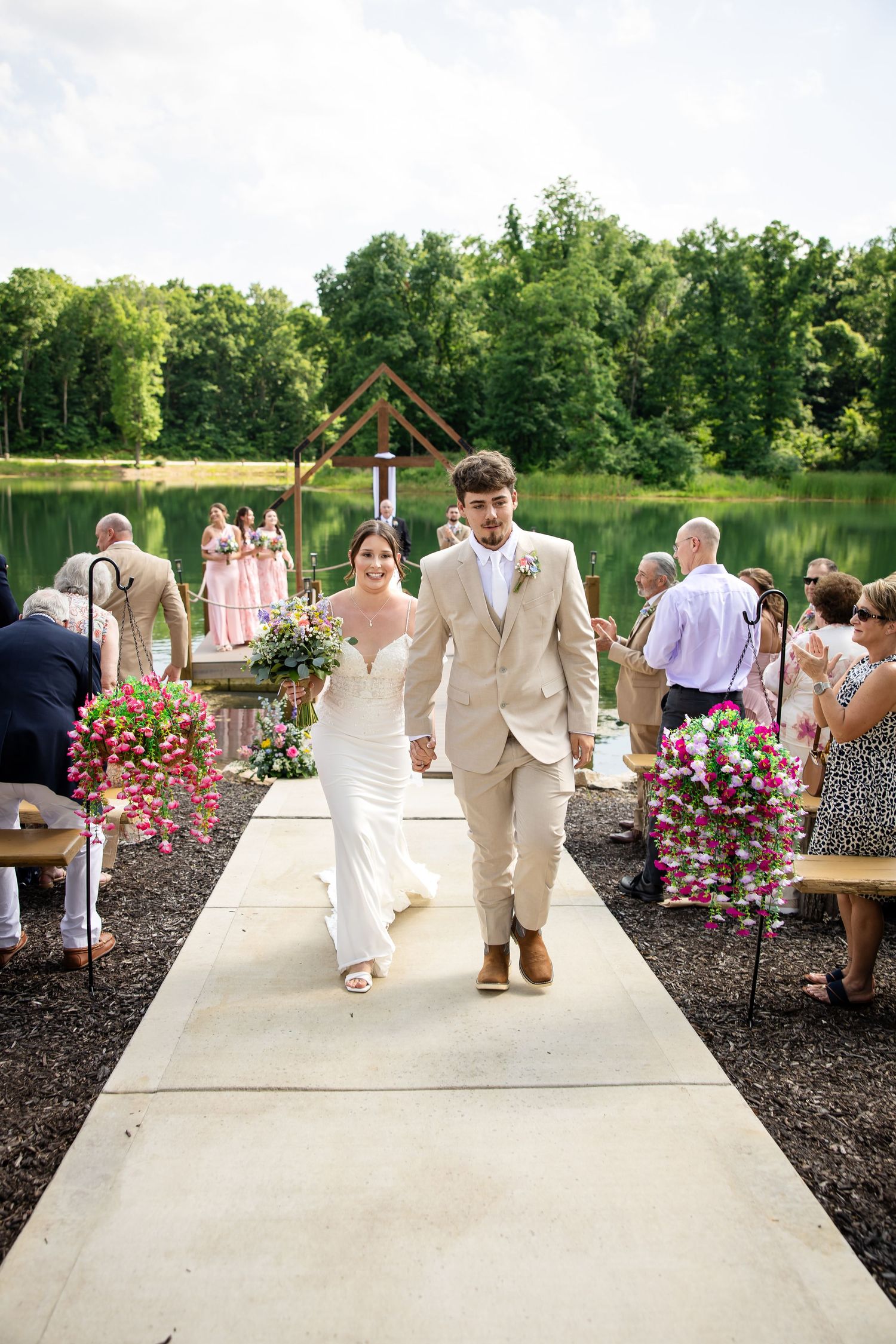 Newlywed couple walks down outdoor wedding aisle by lake, bride in white gown, groom in tan suit, pink floral decor.