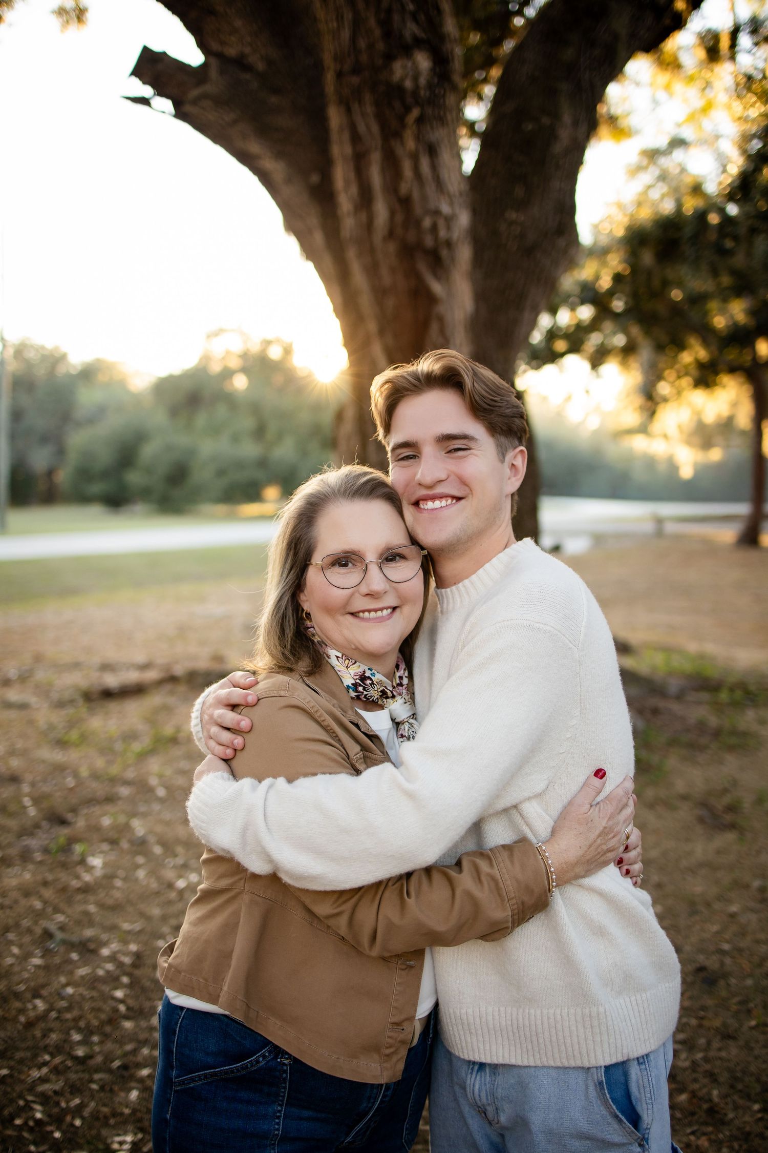 A couple embraces outdoors in casual fall attire with golden sunlight filtering through trees in the background.