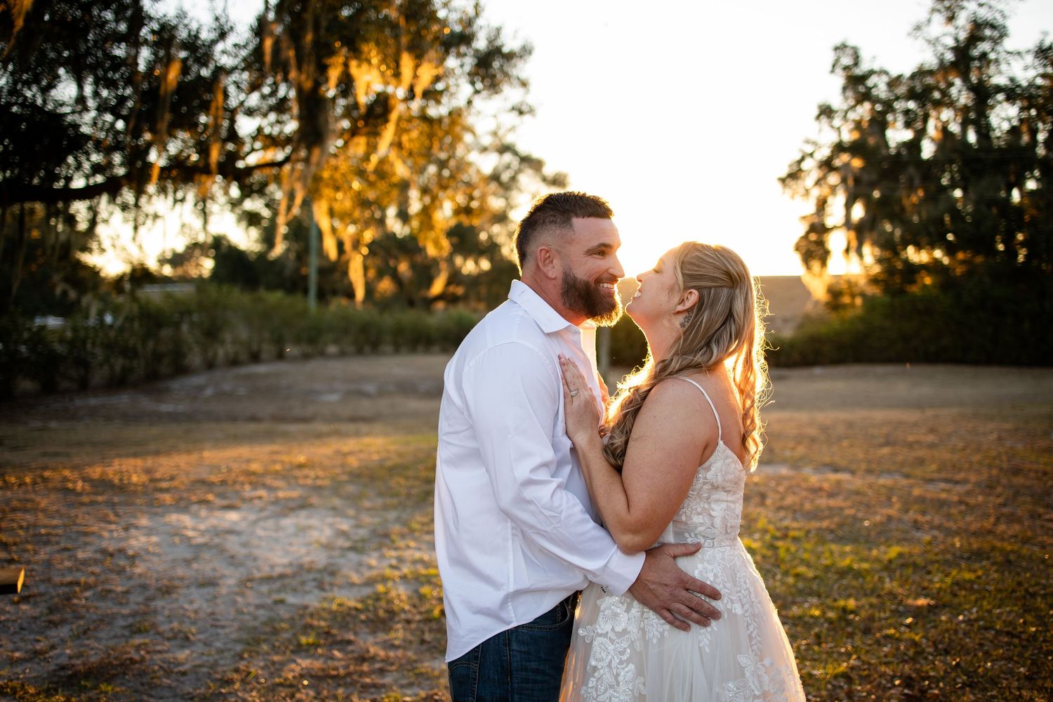 A couple embraces during golden hour sunset in a rustic outdoor setting with Spanish moss hanging from trees.