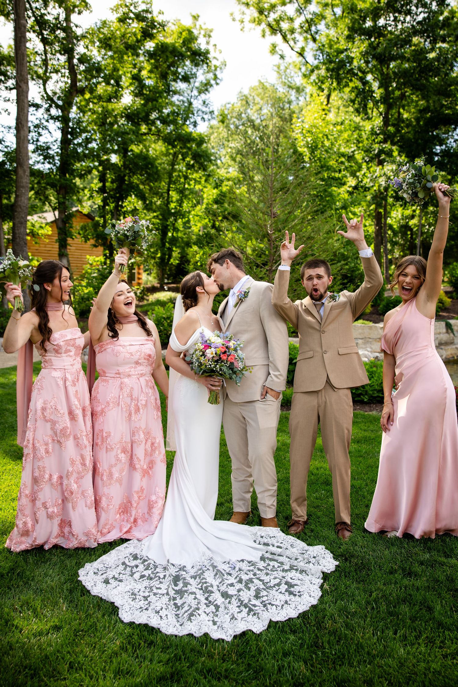 A bride and groom share a kiss surrounded by bridesmaids in pink gowns and a groomsman celebrating outdoors.