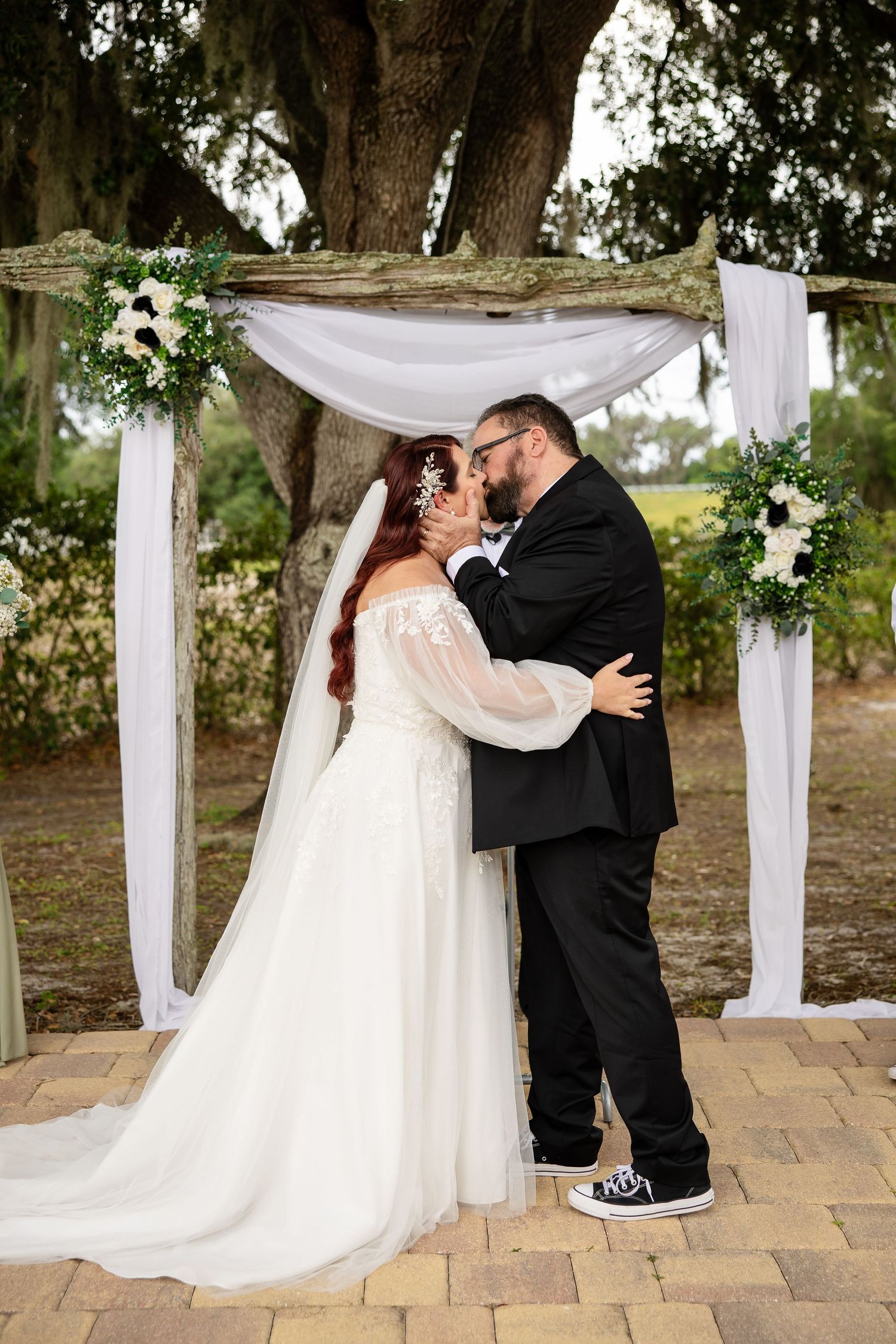 Bride and groom share first kiss under rustic floral arch with white draping outdoors near large oak tree.