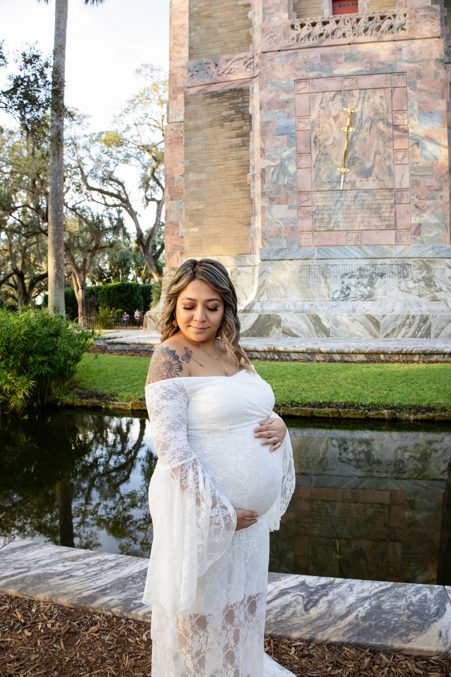 A white lace maternity dress glows in golden sunlight beside a reflective pond near a stone building exterior.