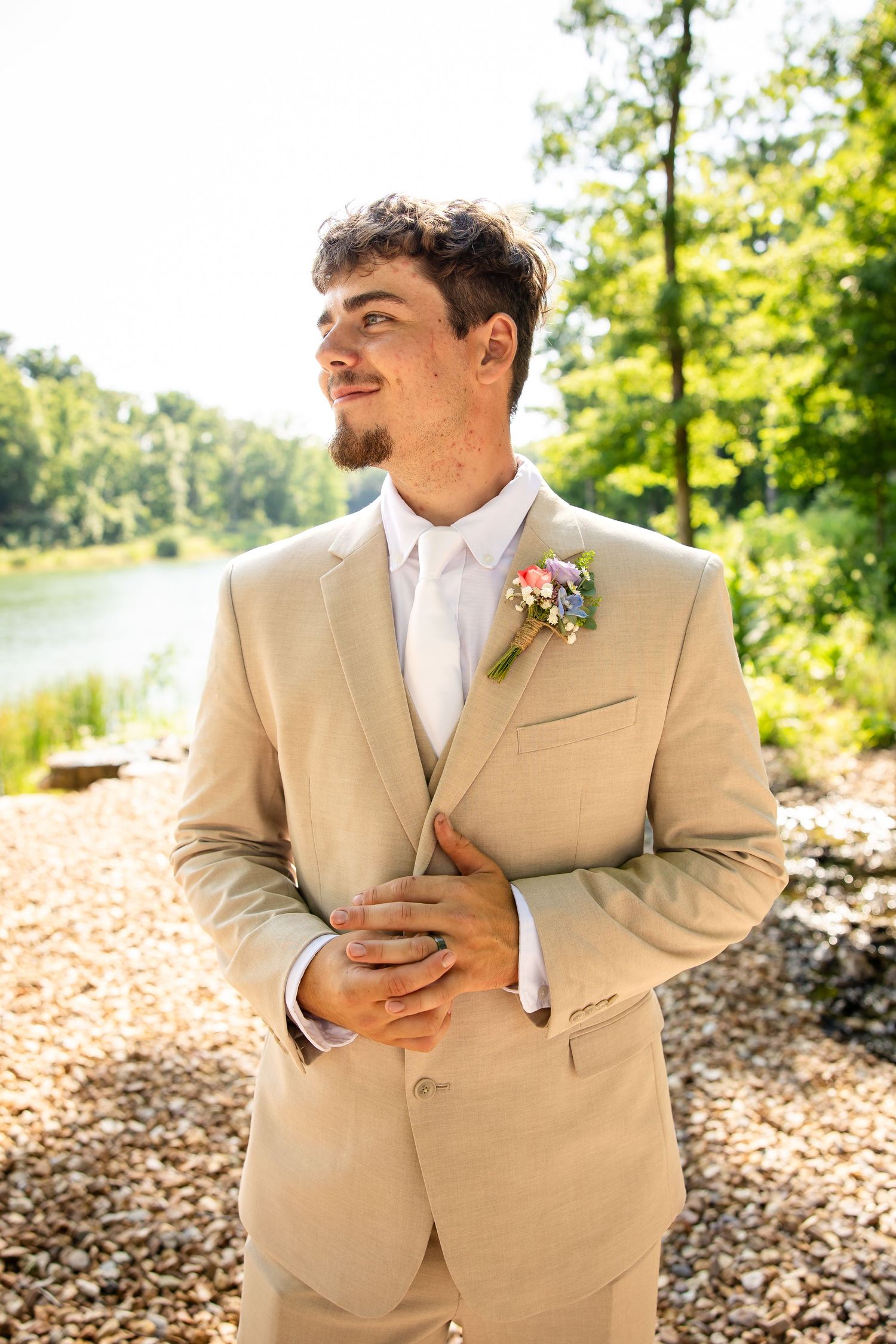 Groom in tan suit with floral boutonniere standing by a river on a rocky shore surrounded by lush green trees.