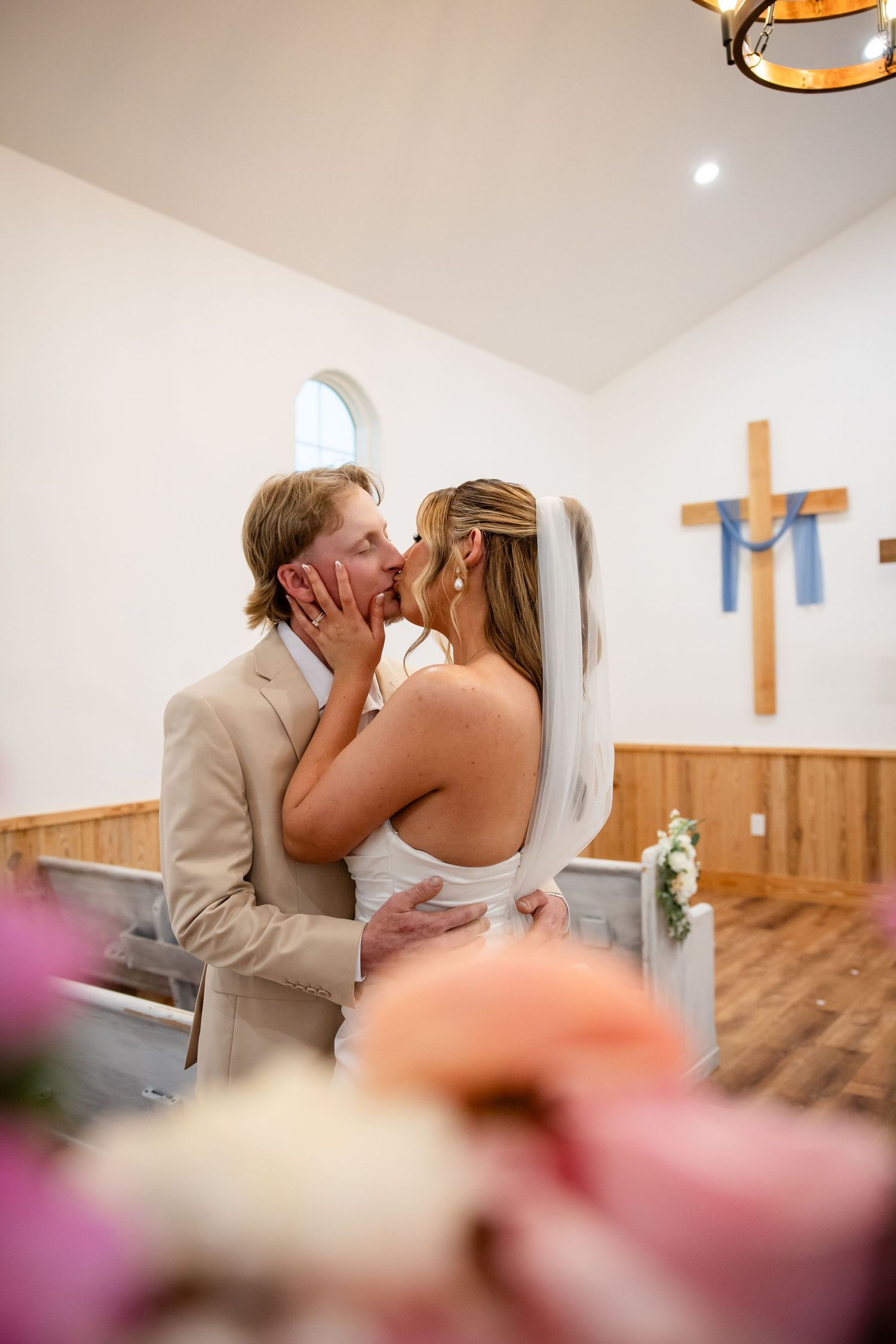 Bride and groom share first kiss at chapel altar, wooden cross on white wall, romantic wedding ceremony moment.