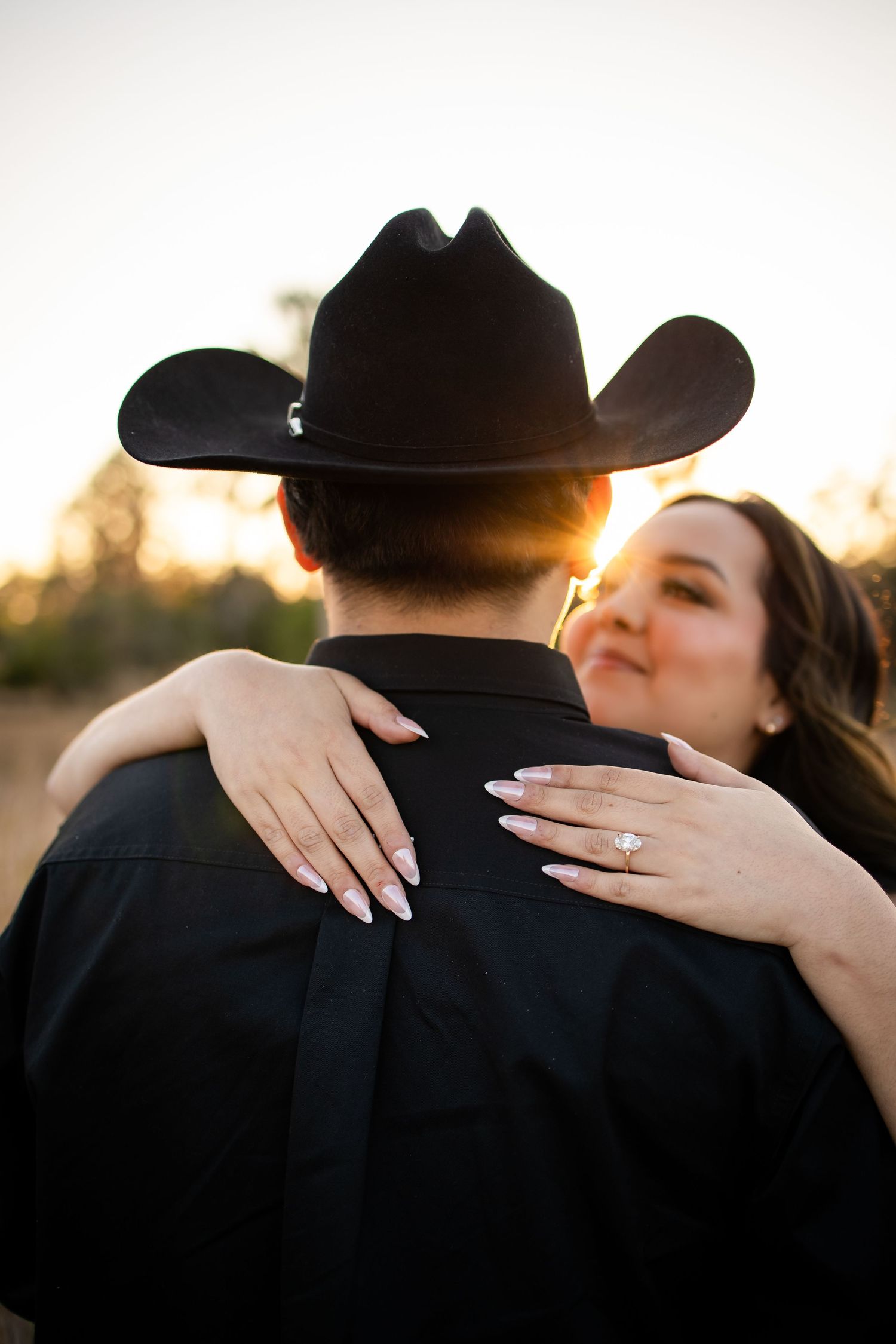 A couple embraces during sunset, with a person wearing a black cowboy hat and shirt while their partner shows off an engagement ring.