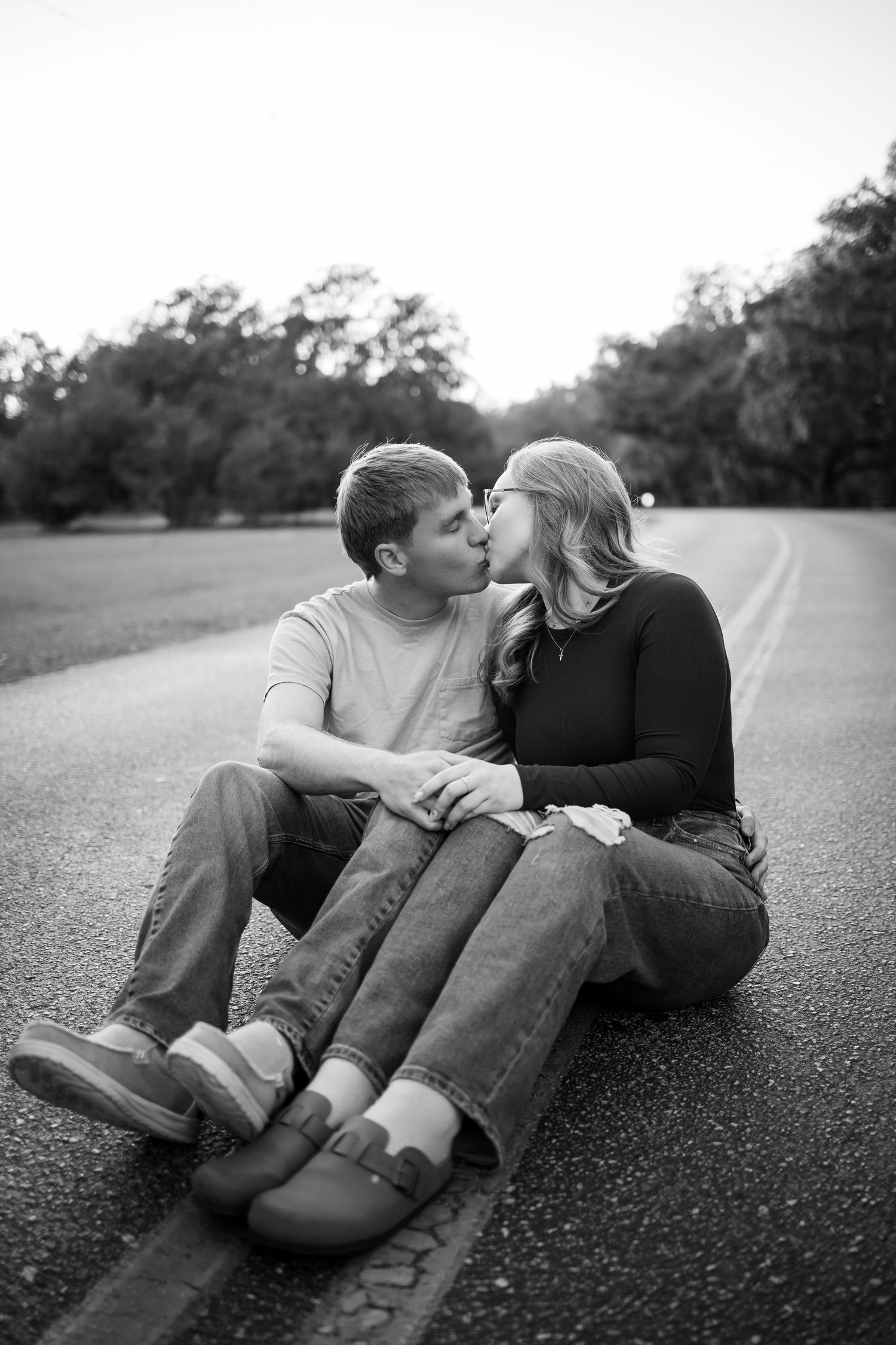 A couple shares a romantic kiss while sitting on an empty road in black and white.