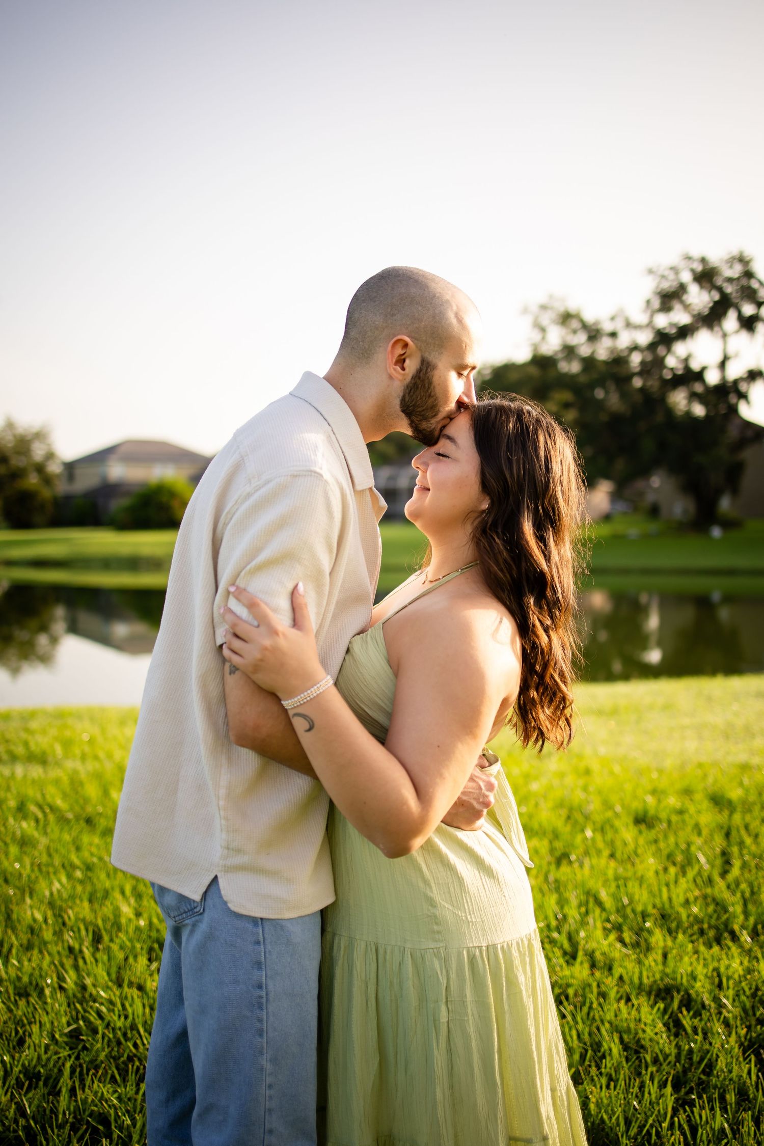 A romantic couple embraces on grassy lawn near a lake at sunset as golden light illuminates their tender moment.