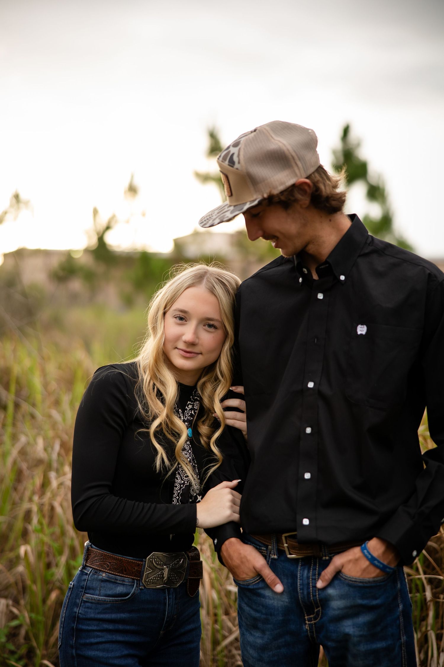 Young couple in black outfits posing outdoors in a field, girl with blonde hair leaning on boy wearing a cap.