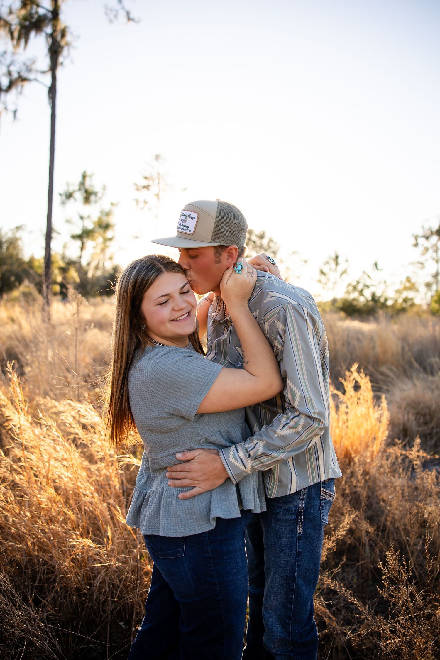 A couple embraces lovingly in a golden field at sunset while wearing casual denim and gray clothing.