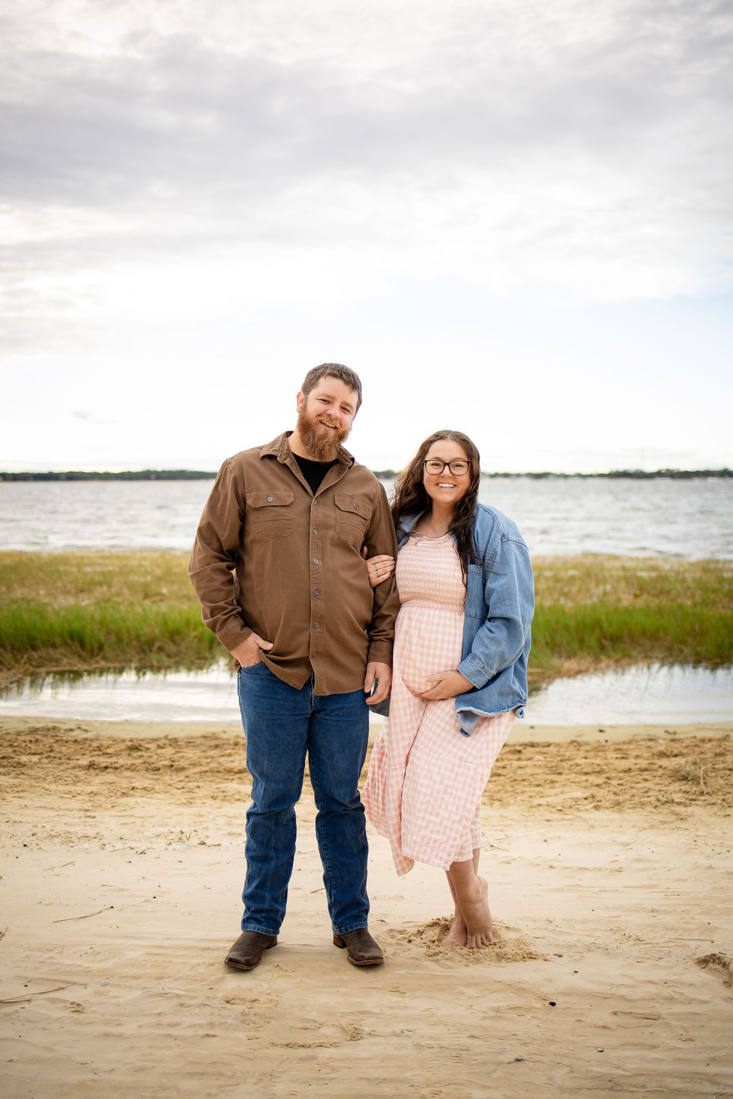 A couple poses together on a beach with grassy marshland and water in the background.