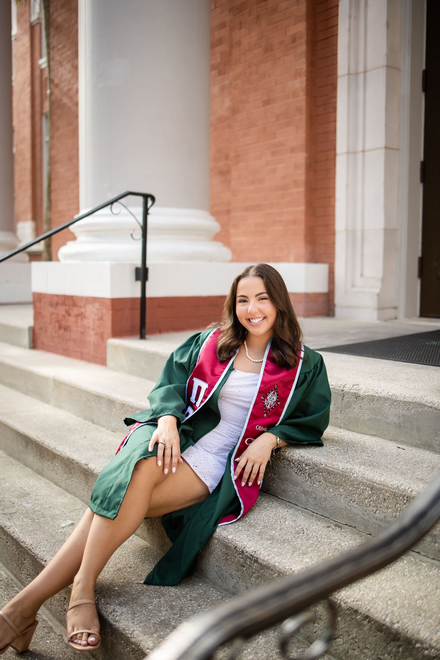 A senior portrait photography session showcases student in green varsity jacket sitting on steps of brick building.