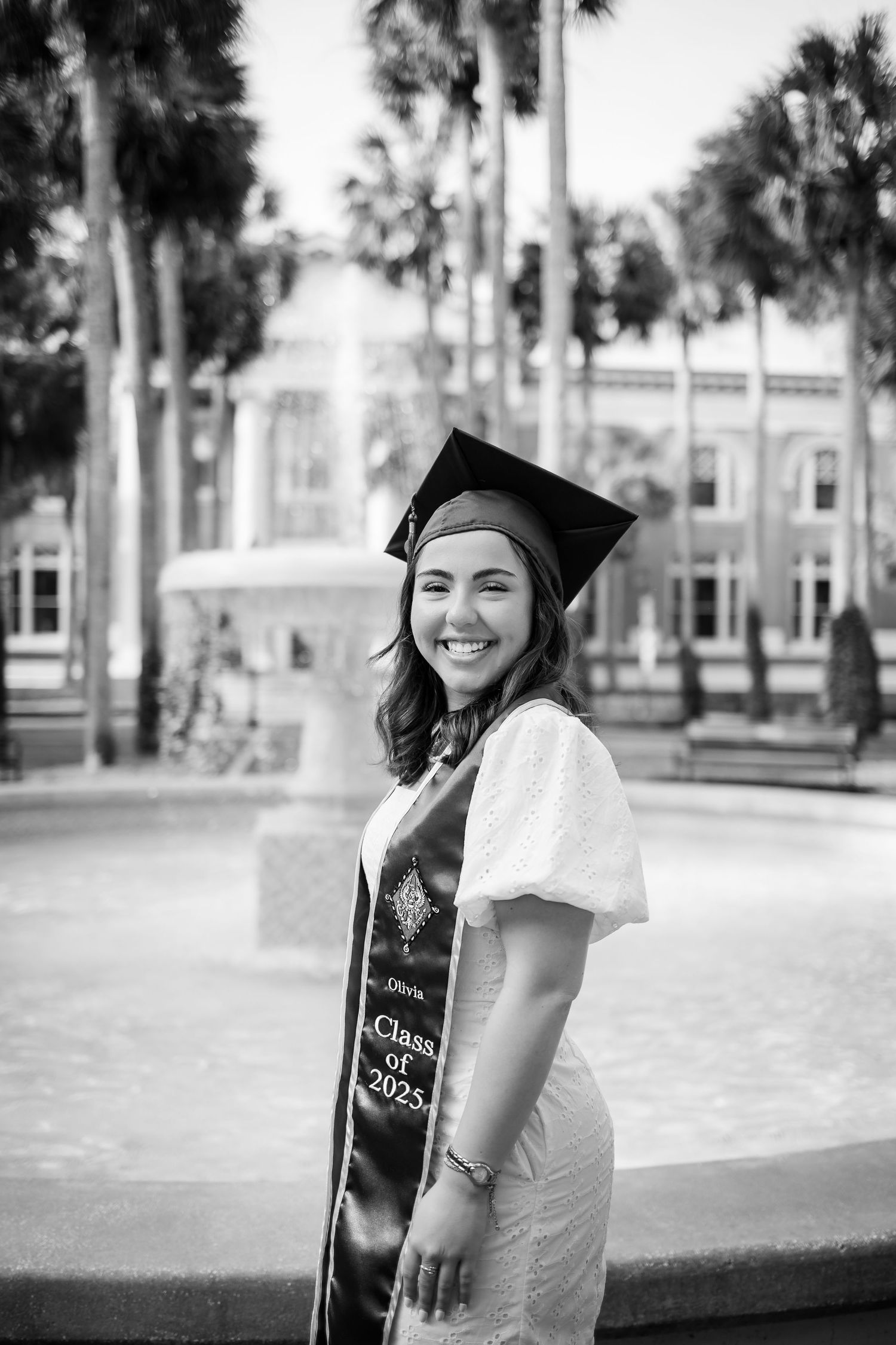 Black and white graduation portrait outdoors with palm trees and a fountain in the background.