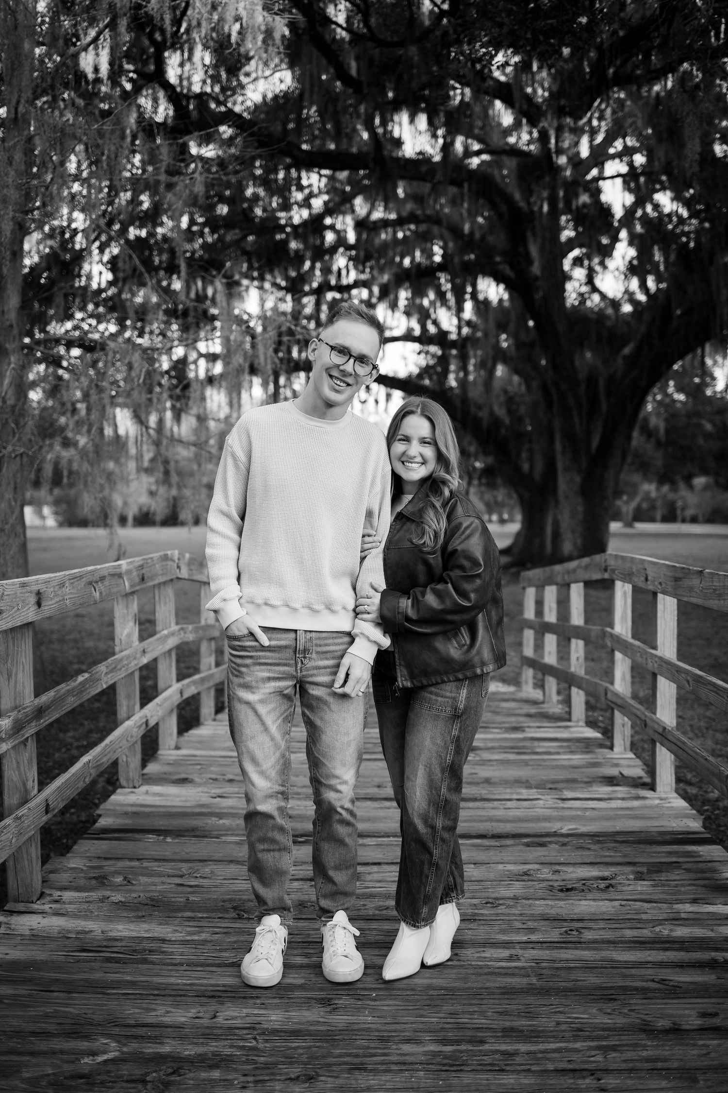 A couple in casual wear stands together on a wooden bridge beneath Spanish moss.