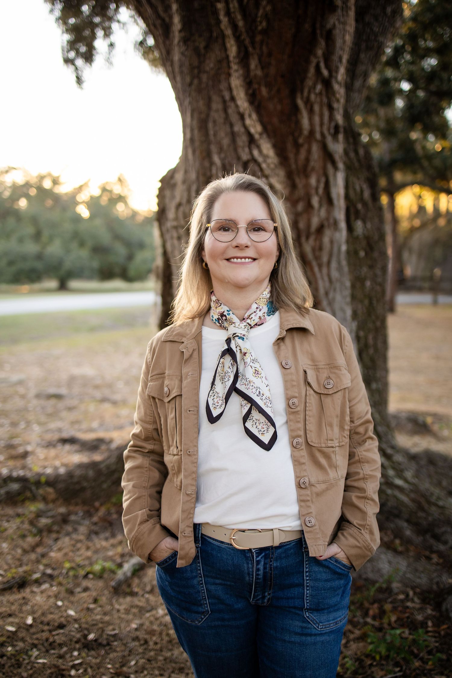 A person in a tan jacket and scarf poses casually with hands in pockets under a large tree.