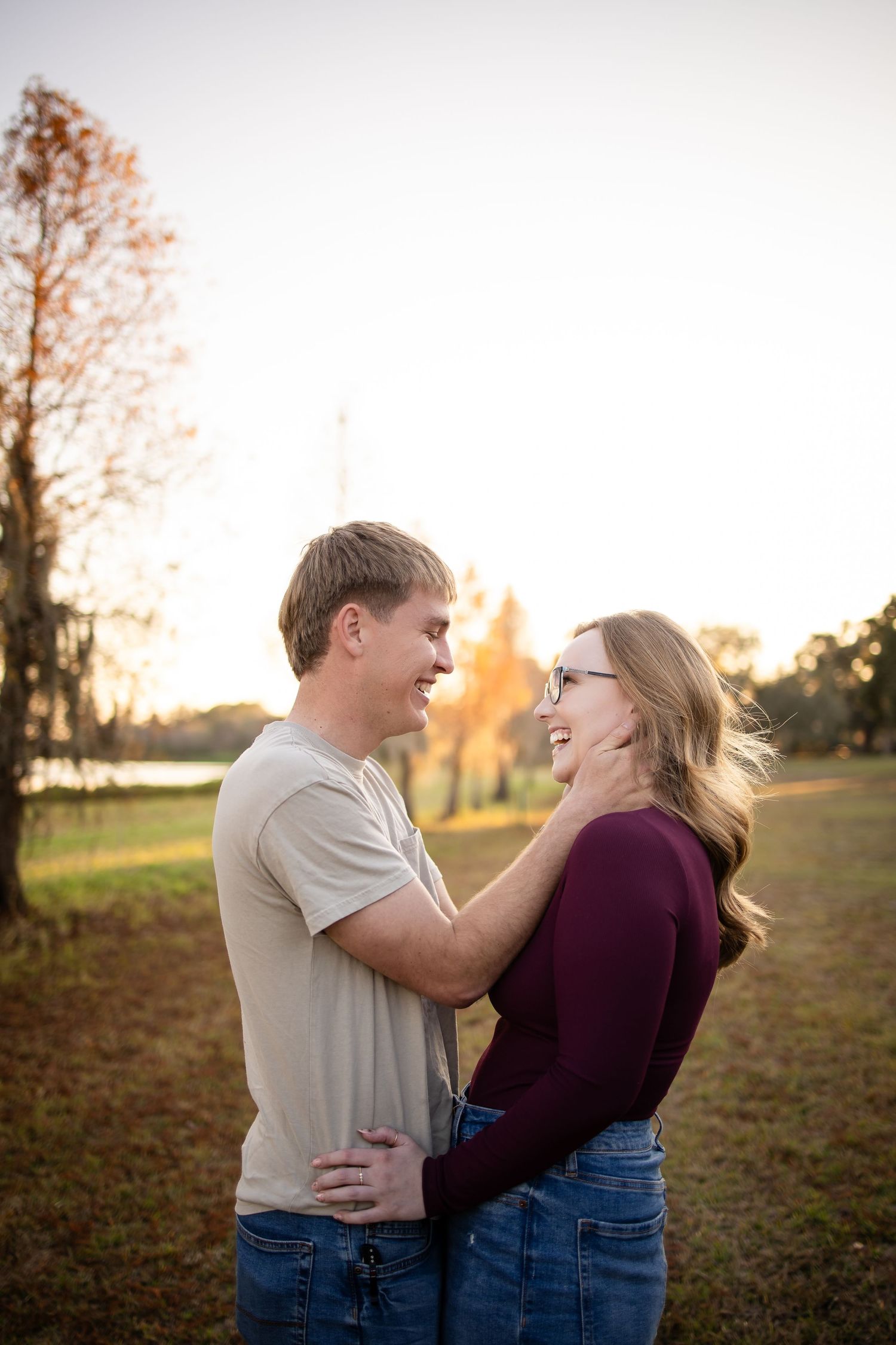 A couple laughs together during golden hour in a park with fall foliage in the background.