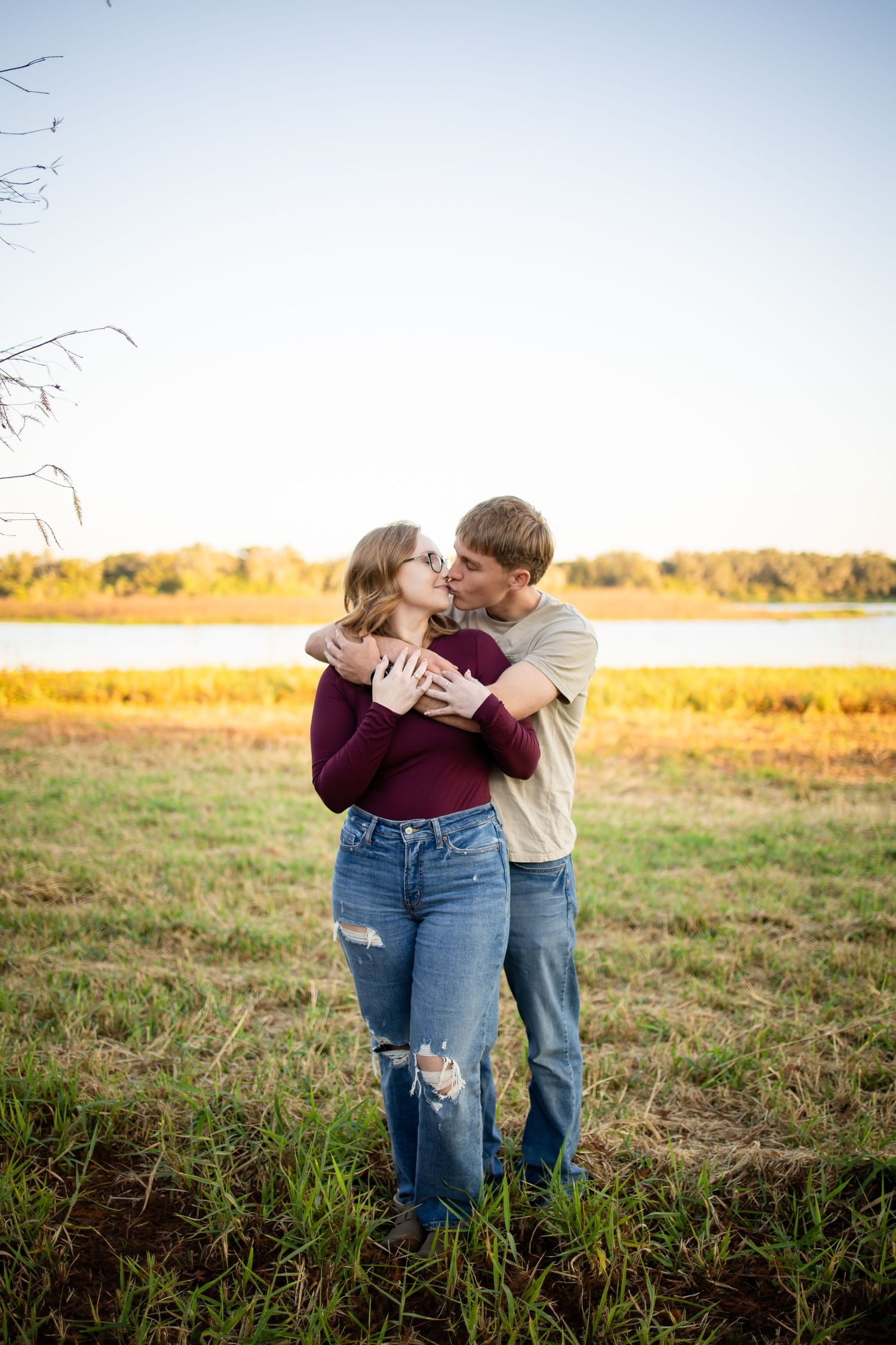 A couple embraces in a golden field during sunset wearing casual denim and burgundy clothing.