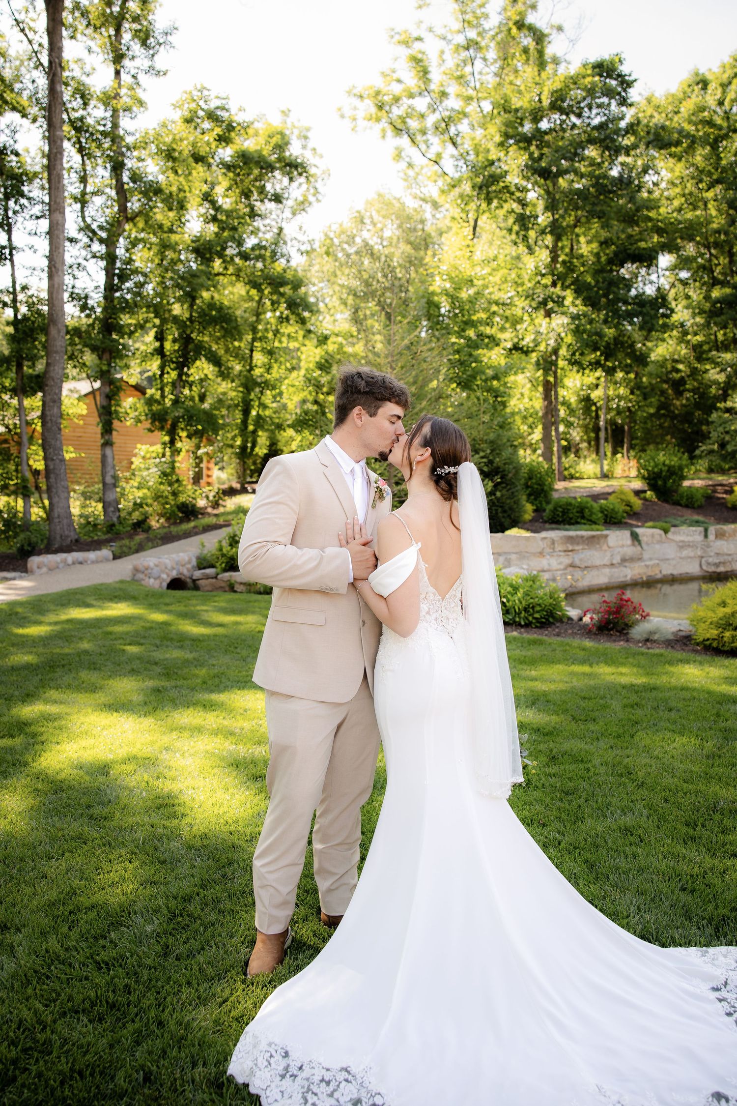 A bride and groom share a romantic moment in a lush green garden, she in a white gown with veil, he in a tan suit.