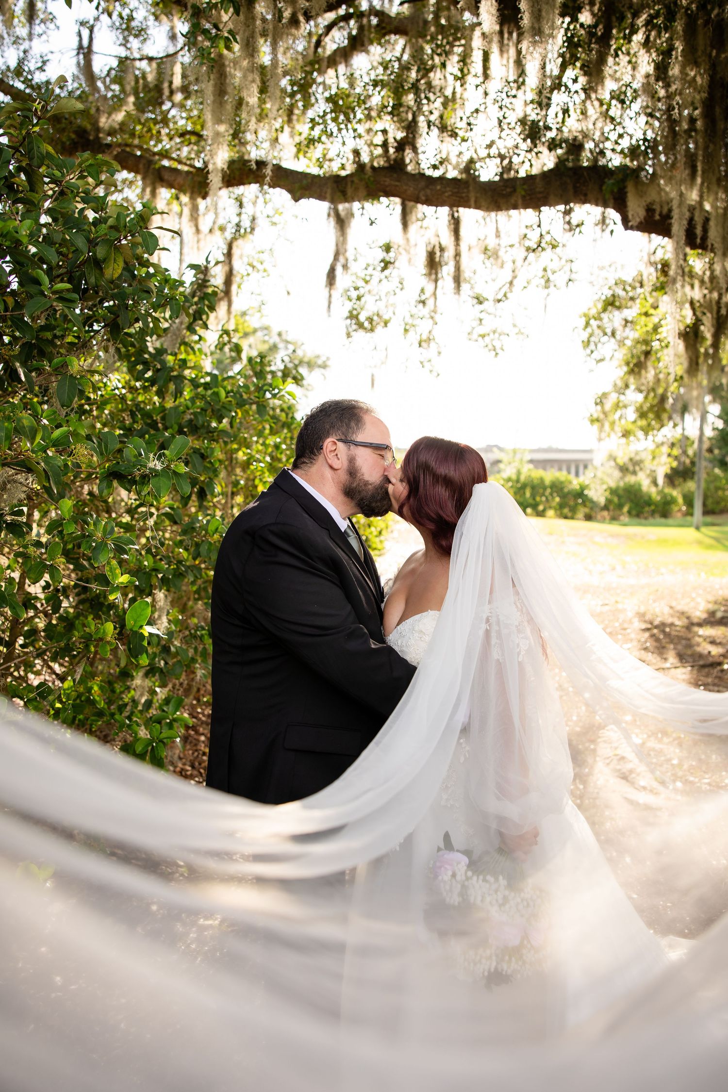 Bride and groom share a romantic kiss under Spanish moss-draped oak tree, with flowing white veil billowing in the breeze.