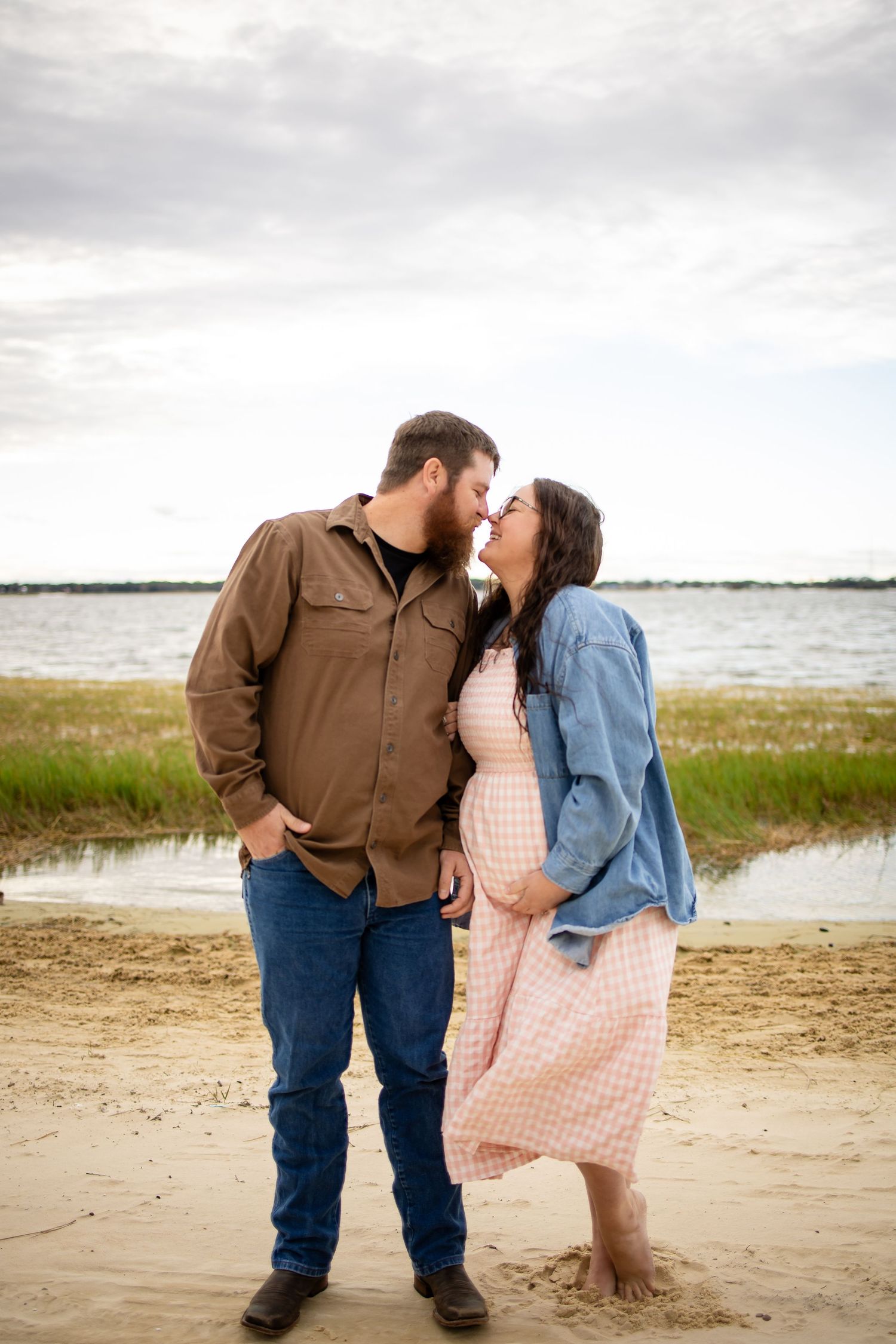 A romantic couple shares an intimate moment on a cloudy beach with wetlands and grass in the background.