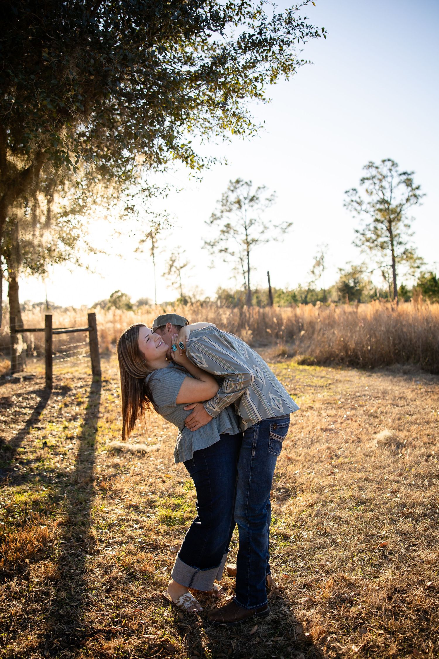A romantic couple embraces in a golden field at sunset with tall trees casting long shadows in the background.