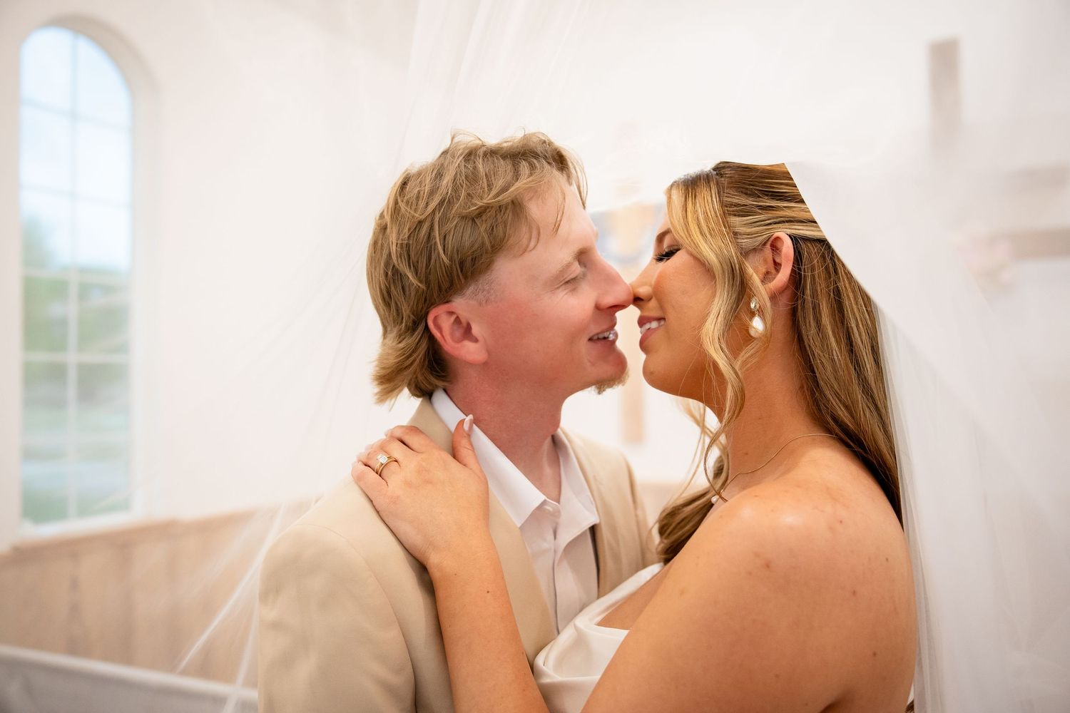 A bride and groom share a romantic moment, nearly kissing, in a softly lit white chapel on their wedding day.