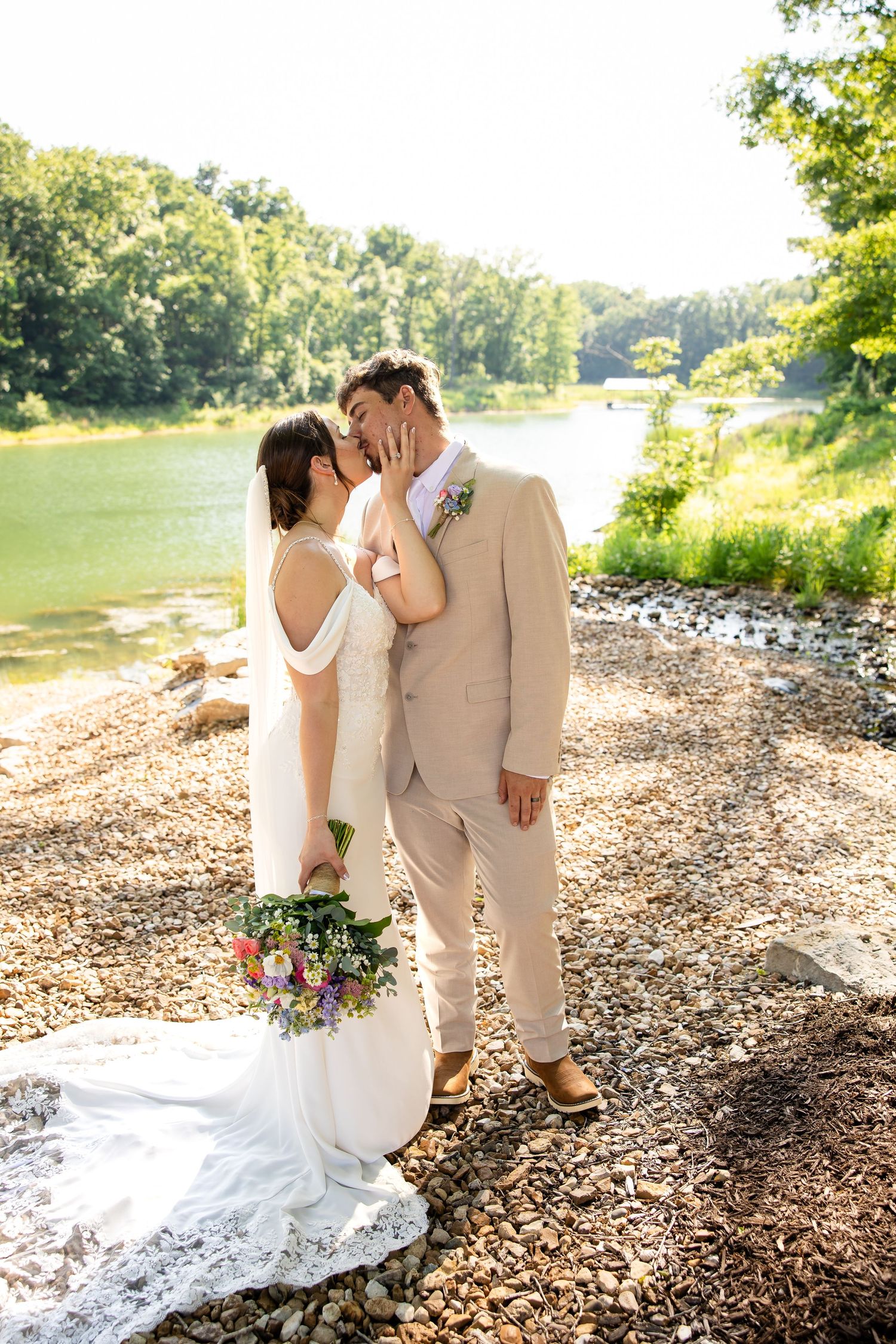 Bride and groom share a romantic kiss by a river, she holds a wildflower bouquet, he wears a tan suit.