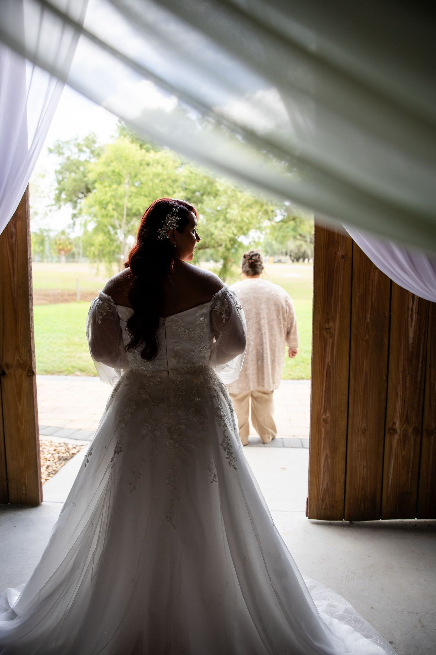 Bride in white lace off-shoulder gown stands in barn doorway as another person walks outside on wedding day.