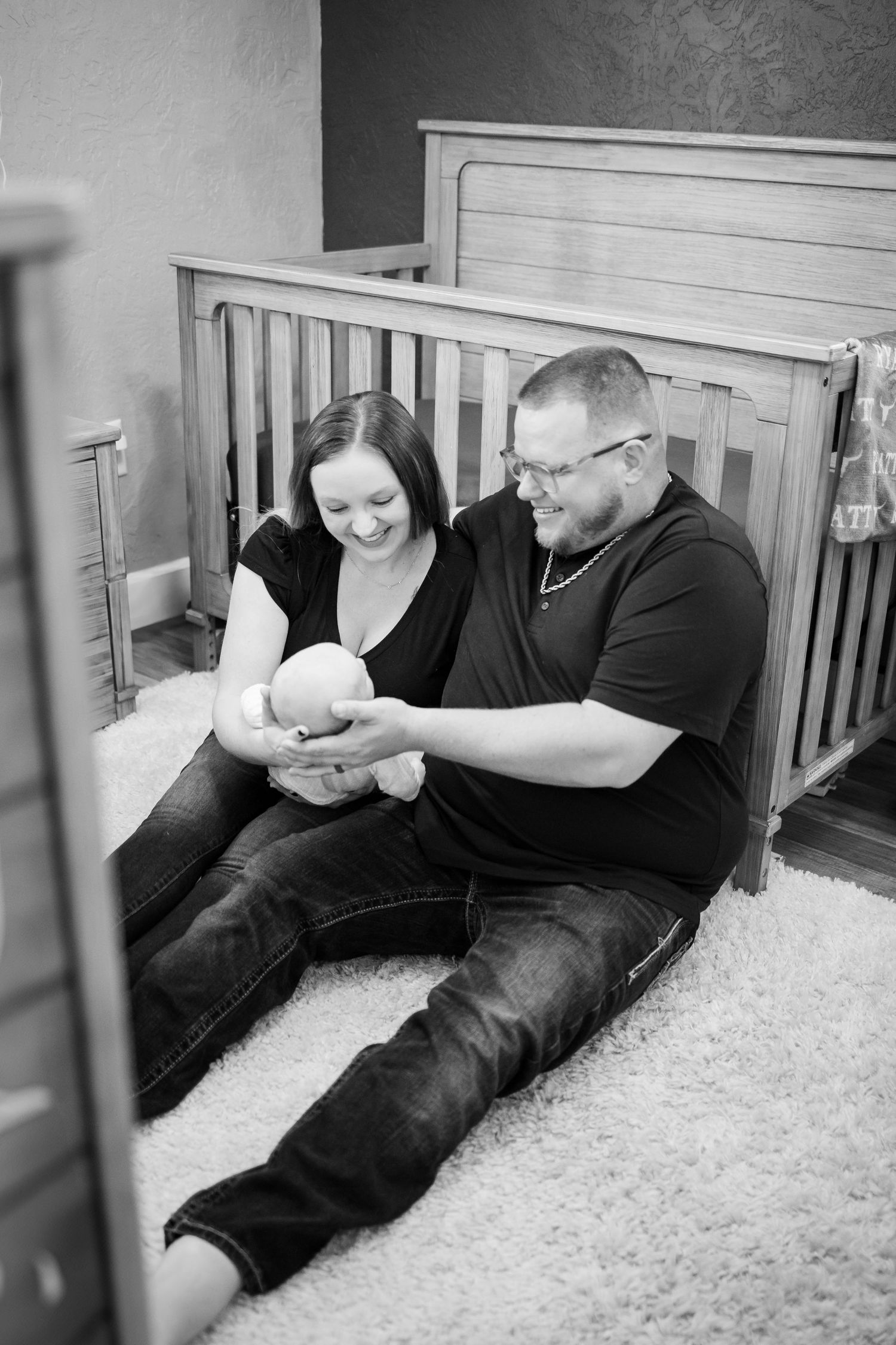 A couple sits on carpet beside a wooden crib sharing a tender moment with their baby.