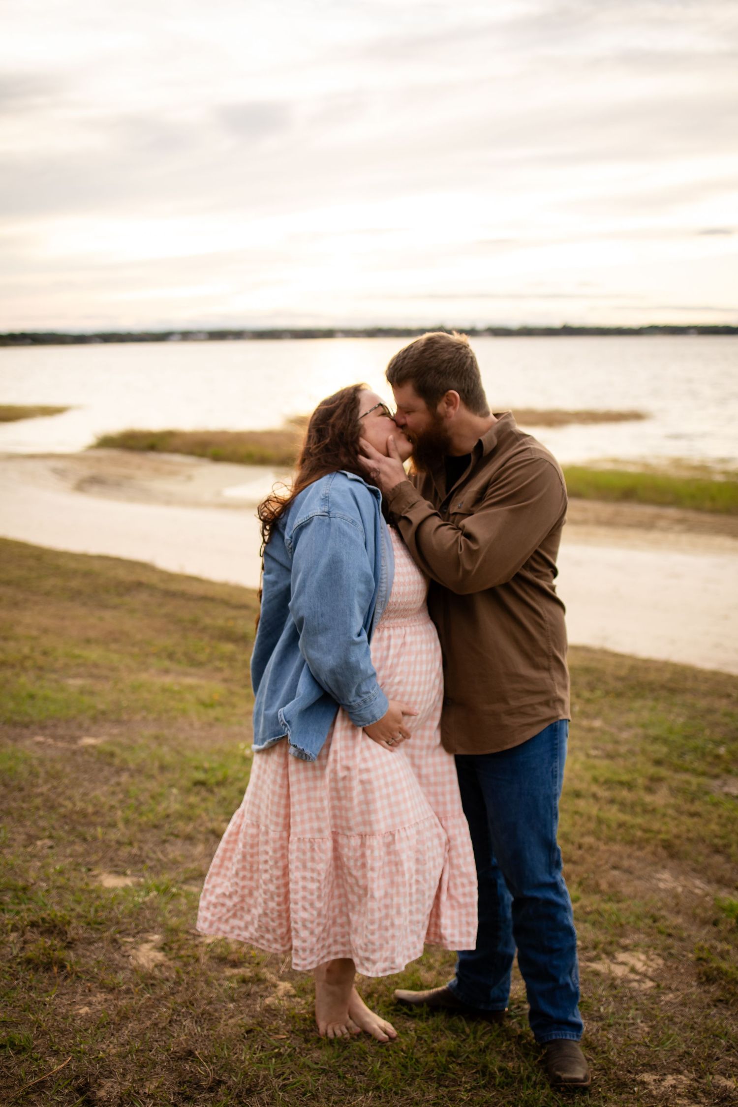 A romantic moment captured at sunset with a couple in denim and brown clothing on grassy shoreline.
