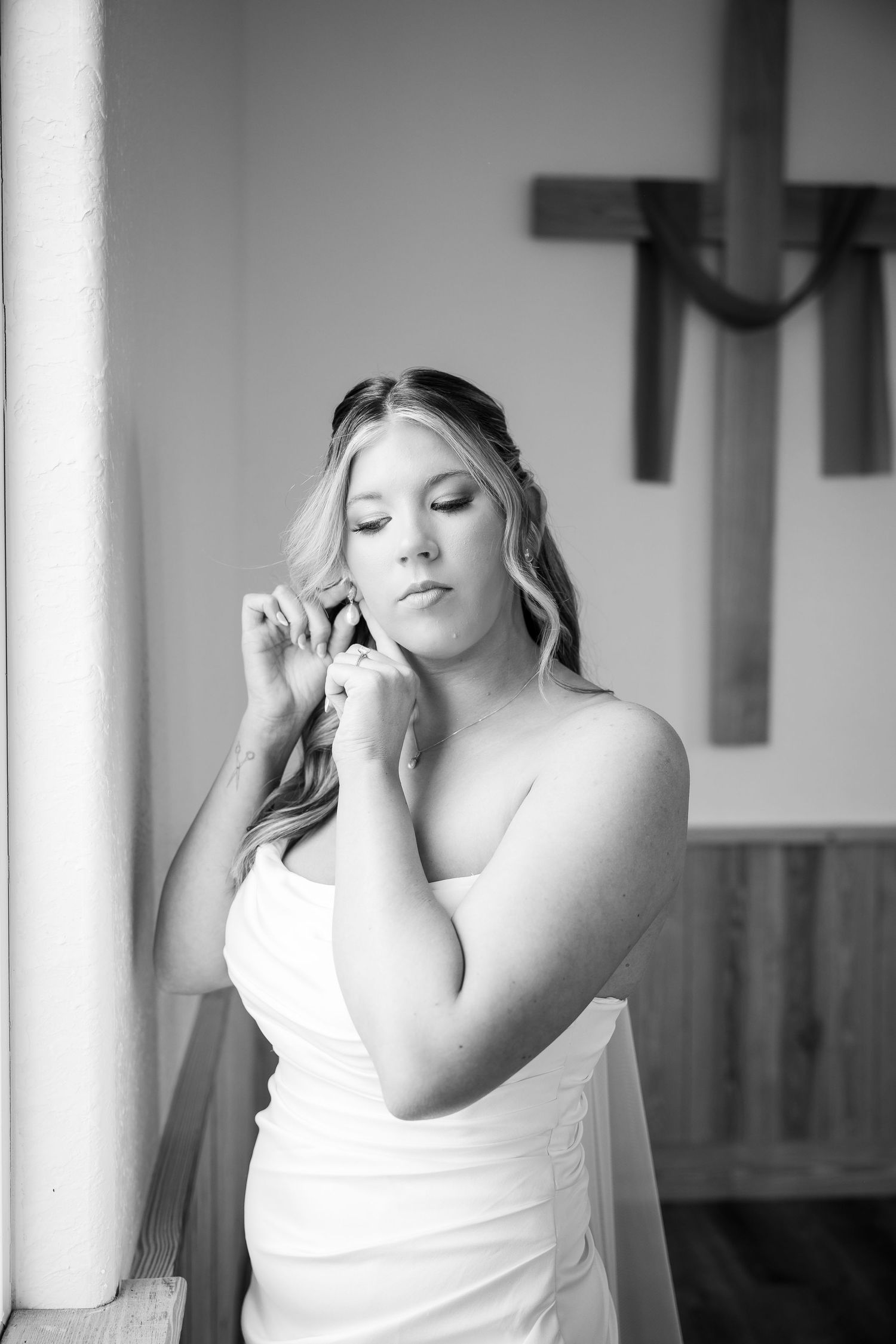 Black and white photo of a bride in a strapless gown adjusting her earring near a wooden cross in a church.