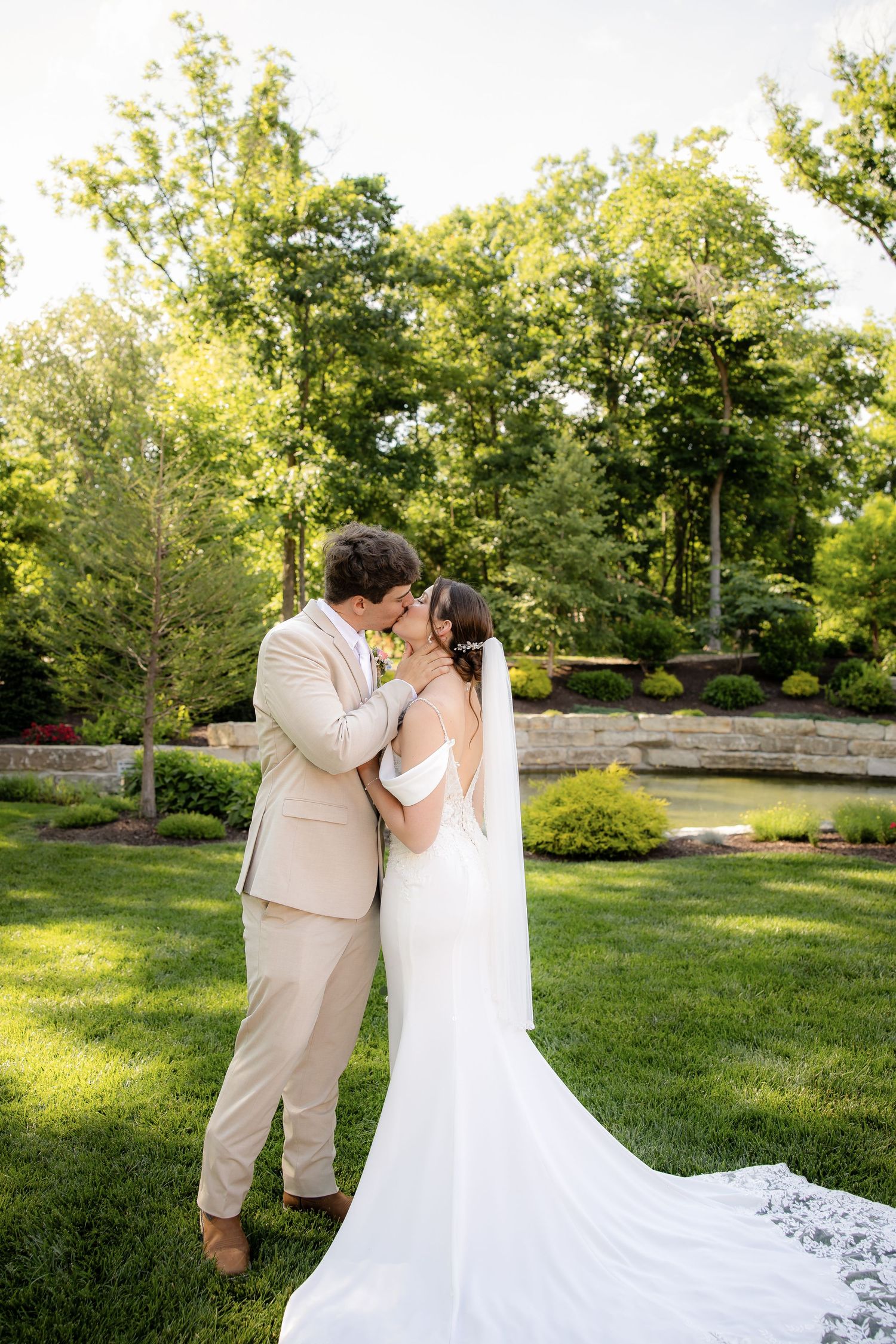 A bride and groom share a romantic kiss in a lush green garden, she wearing a flowing white gown with veil.