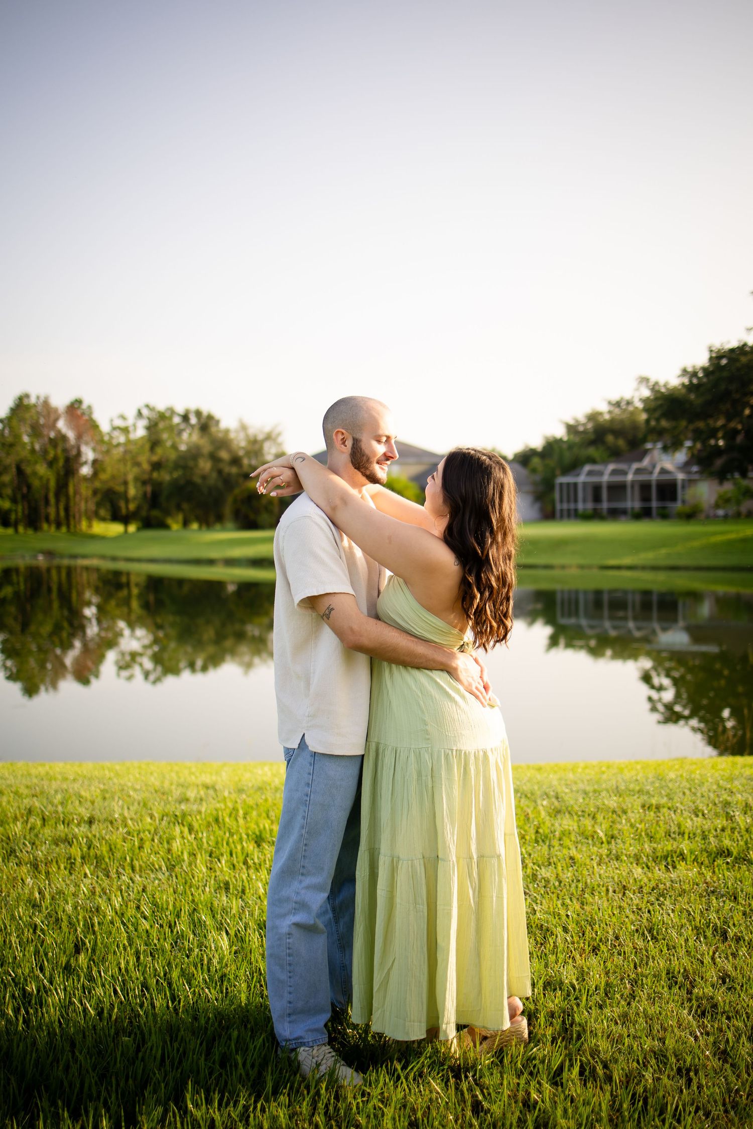 A couple embraces by a serene lake at sunset, with the woman wearing a flowing mint green dress on lush grass.