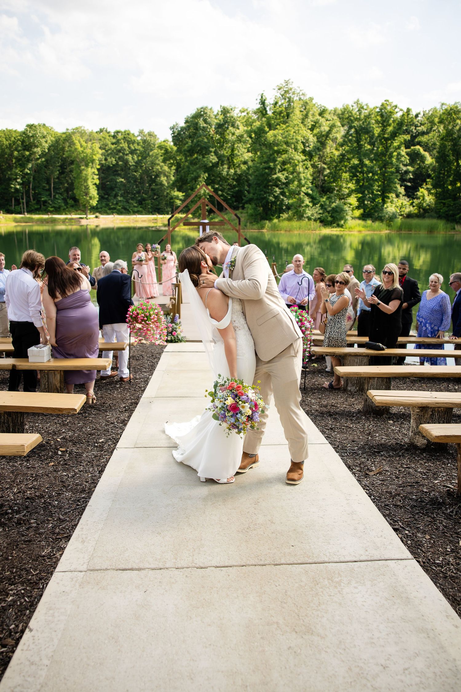 Newlyweds share a kiss on the aisle after outdoor lakeside wedding ceremony with guests watching.
