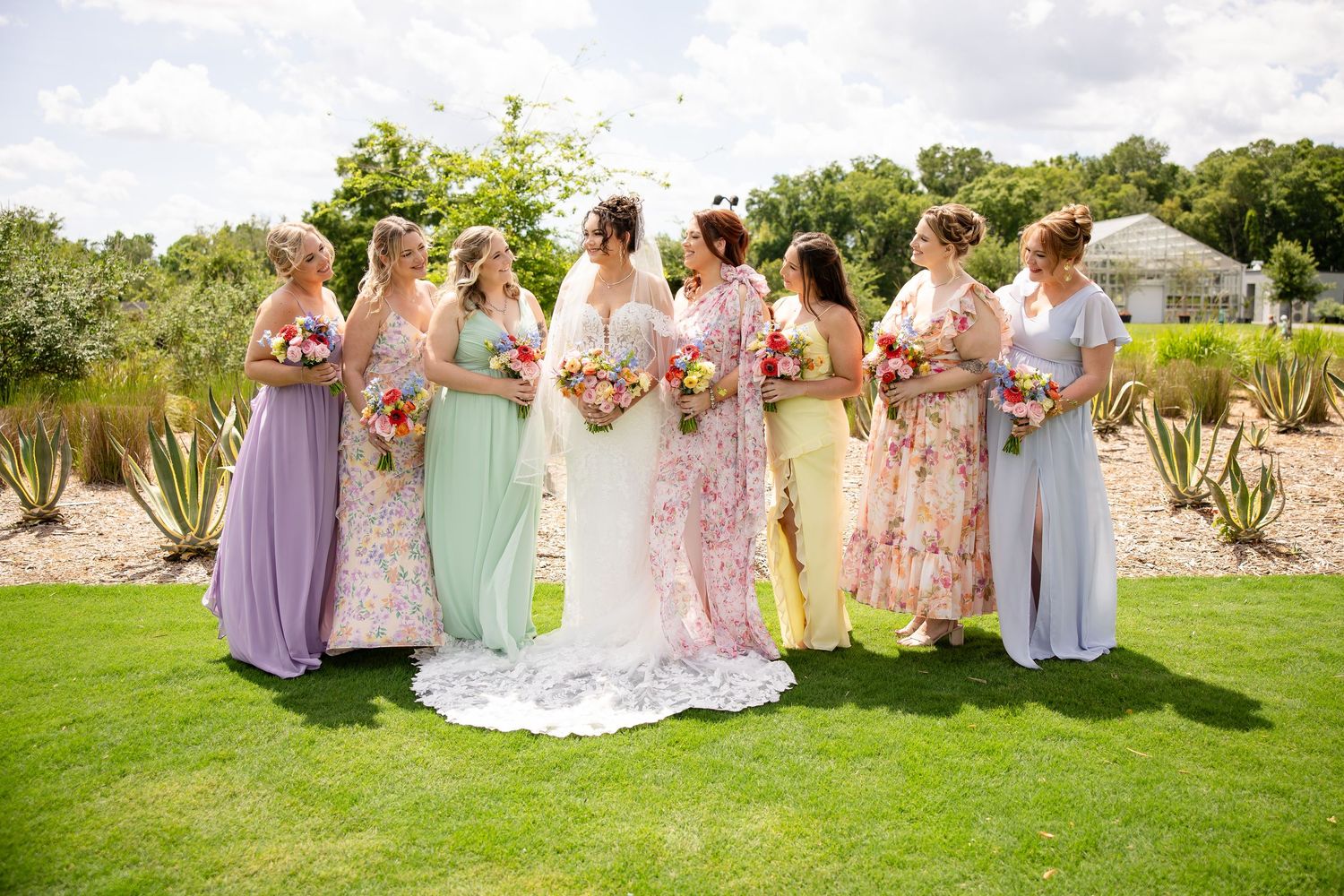 Bride and bridesmaids in pastel dresses holding colorful bouquets on a sunny green lawn at an outdoor wedding.