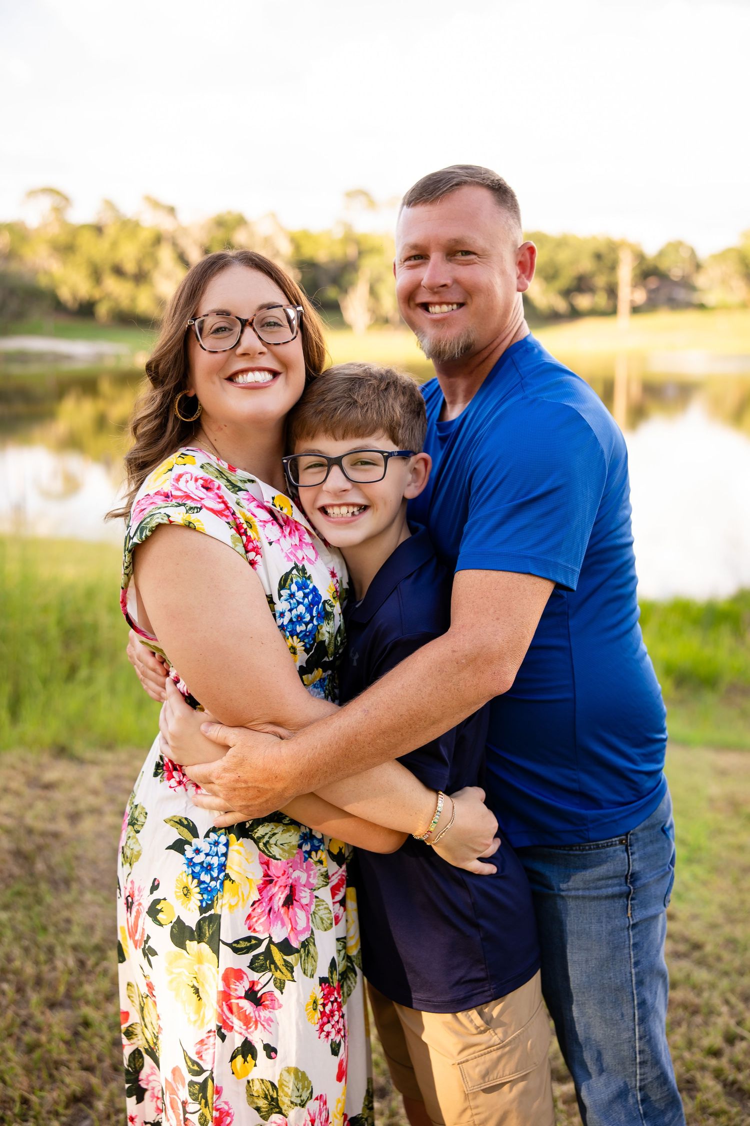 A smiling family of three hugging outdoors near a pond, with a woman in floral dress, boy in navy, and man in blue polo.