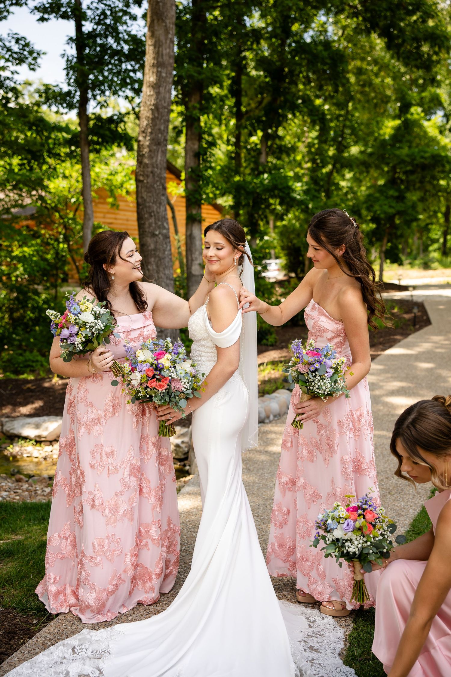 Bride in white gown with two bridesmaids in pink floral dresses holding colorful bouquets outdoors.