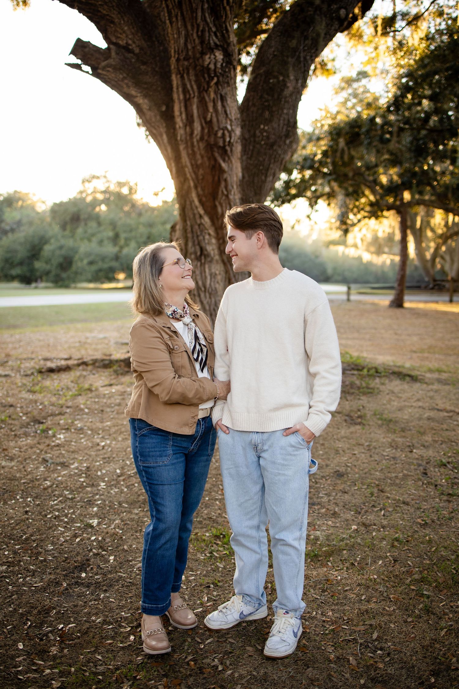 Couple in casual wear shares intimate moment under large tree at golden hour.