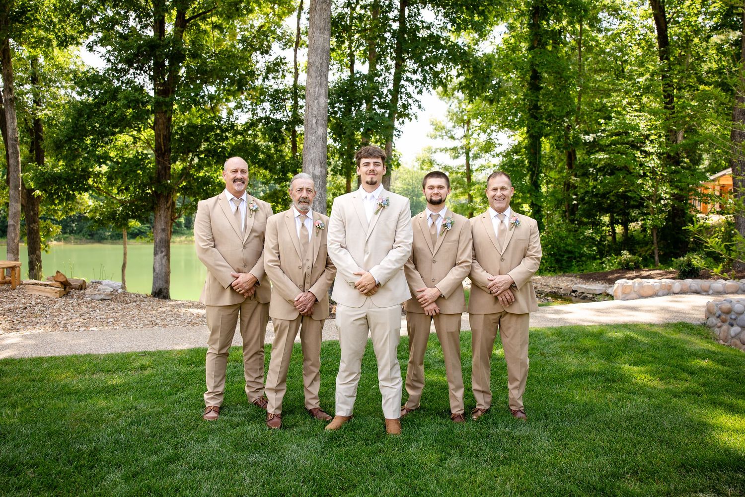 Groom and groomsmen in tan suits posing outdoors on green lawn with lush trees in background at wedding.