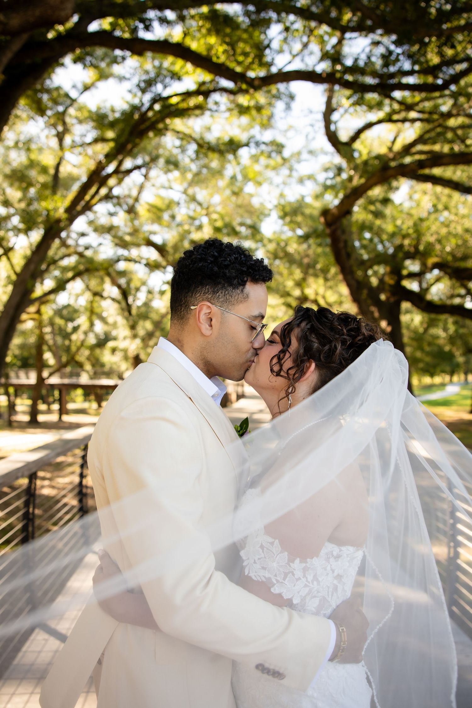 Bride and groom share a kiss under oak trees, her lace veil flowing around them, groom in beige suit.