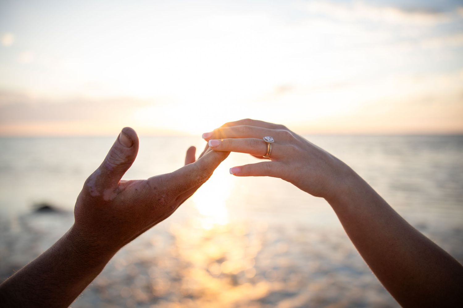 Two hands reaching toward each other at sunset on a beach, one wearing a diamond engagement ring.